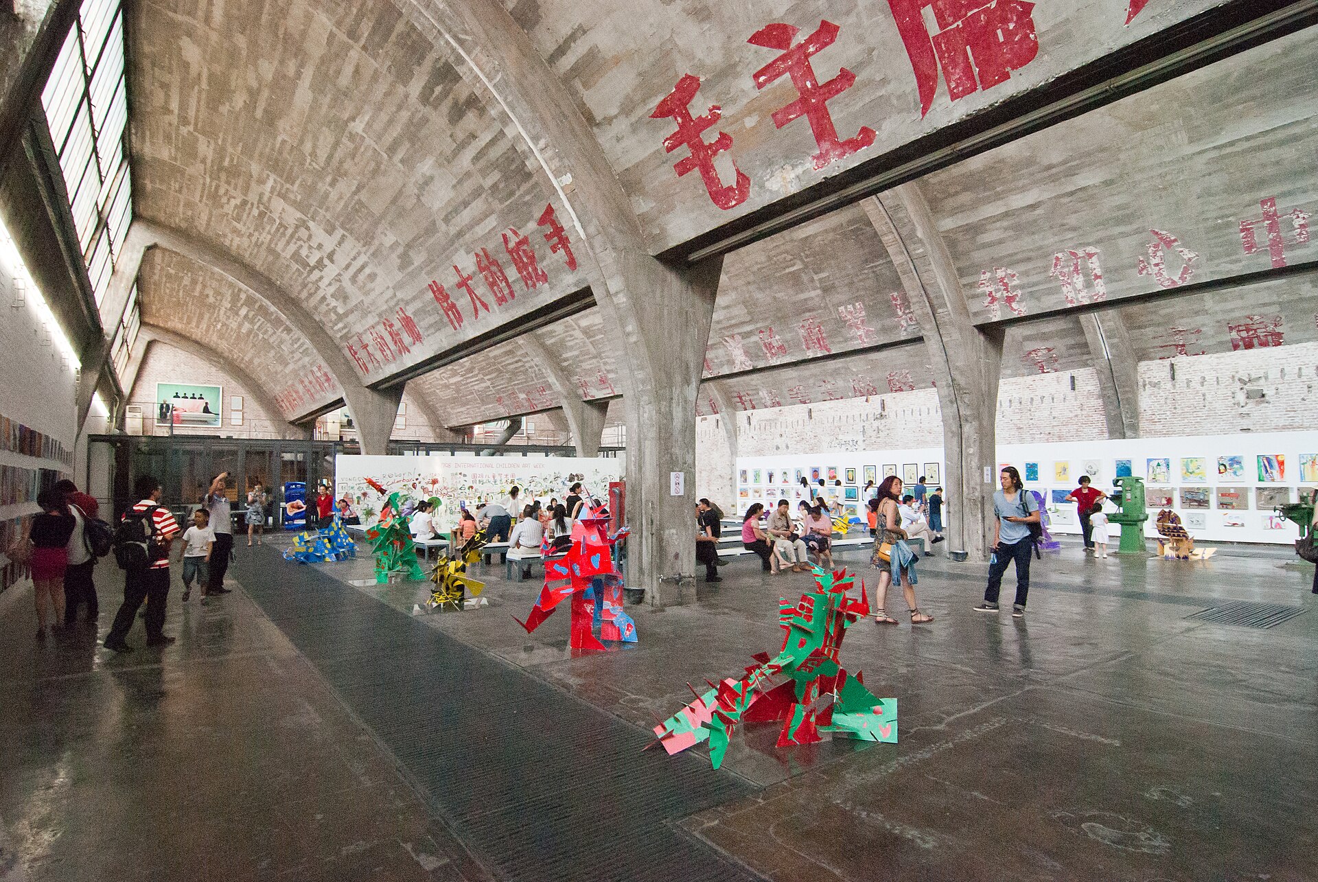 Visitors in a vaulted concrete factory hall at 798 with painted red Chinese slogans overhead and sculptures on the floor