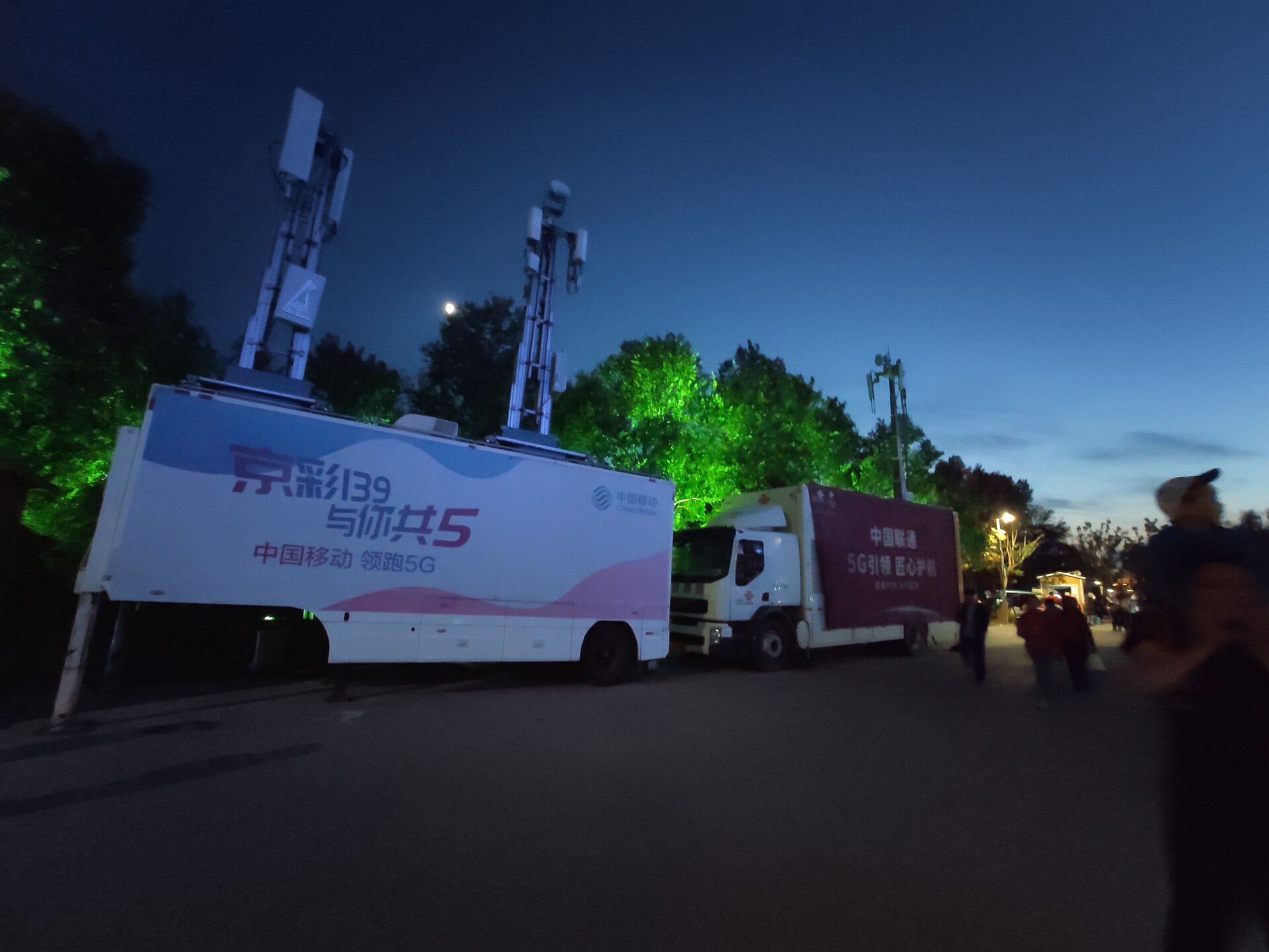 Two mobile 5G base-station trucks from China Mobile and China Unicom with antenna masts, parked at dusk at the 2019 Beijing Expo