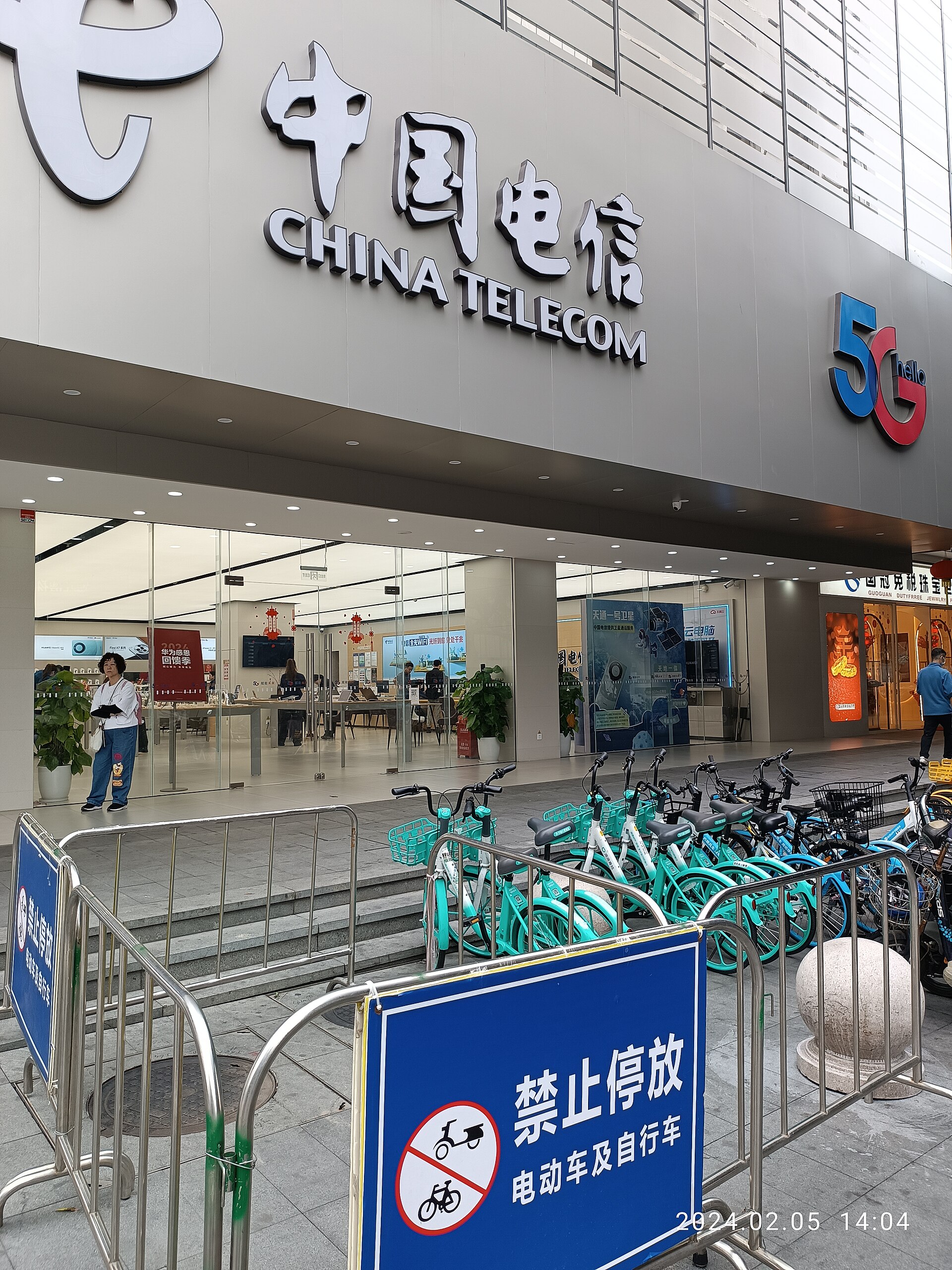 The glass frontage of a large China Telecom store with a 5G sign, and a row of teal shared bicycles parked outside