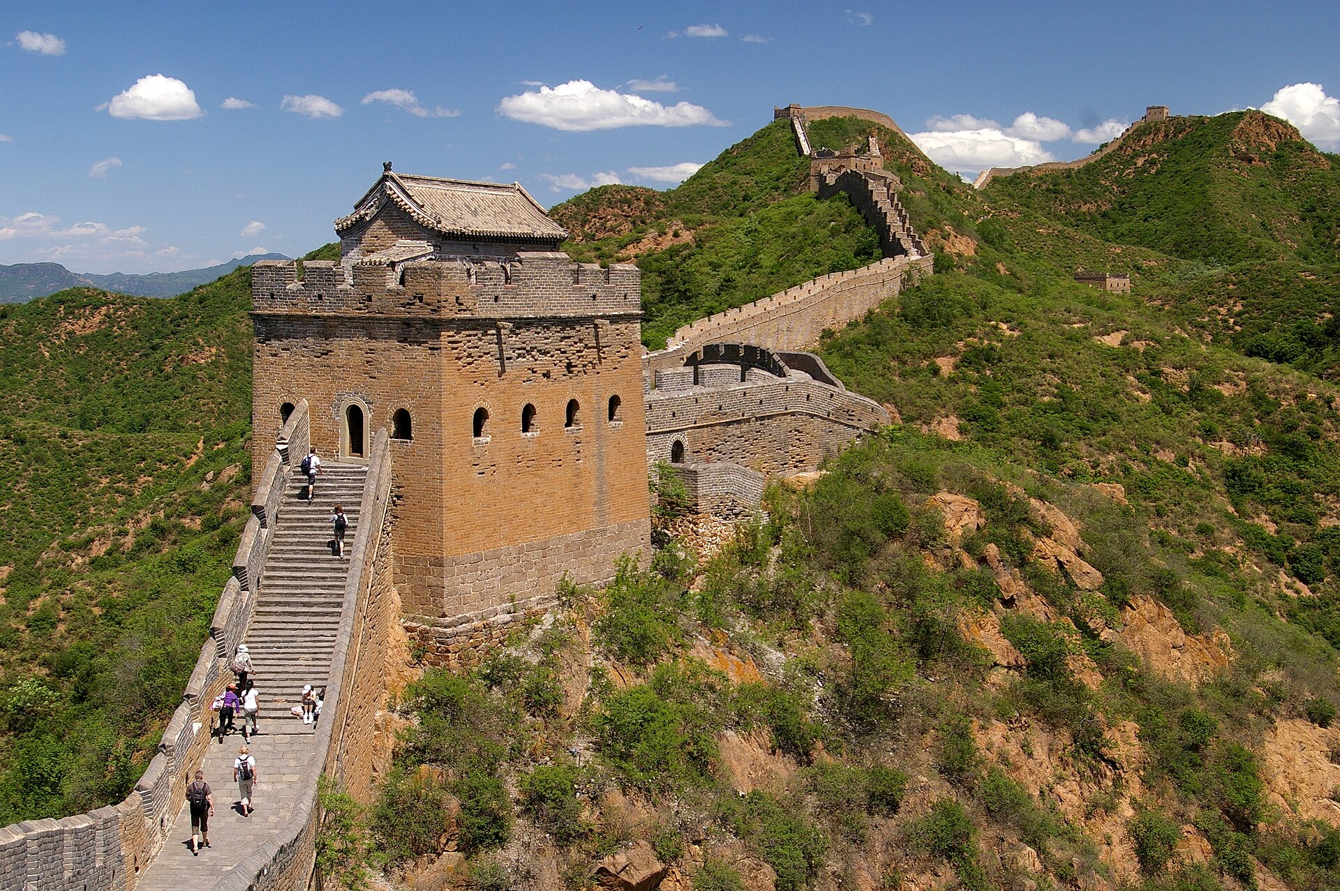 A large brick watchtower on the Jinshanling Great Wall with the rampart running away over green ridges and a few walkers on the steps