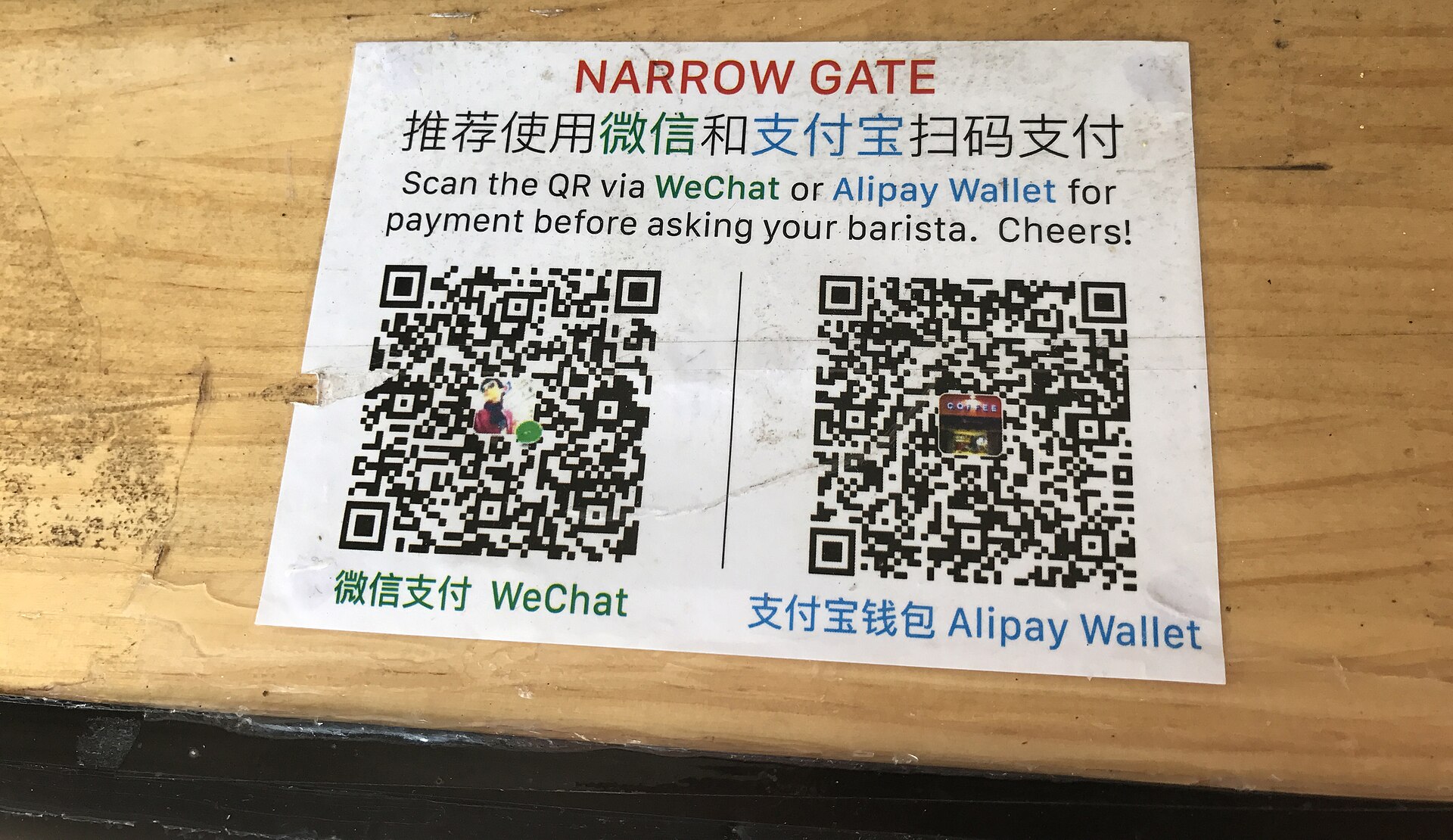 A printed card on a wooden café counter in China showing WeChat and Alipay QR codes for payment