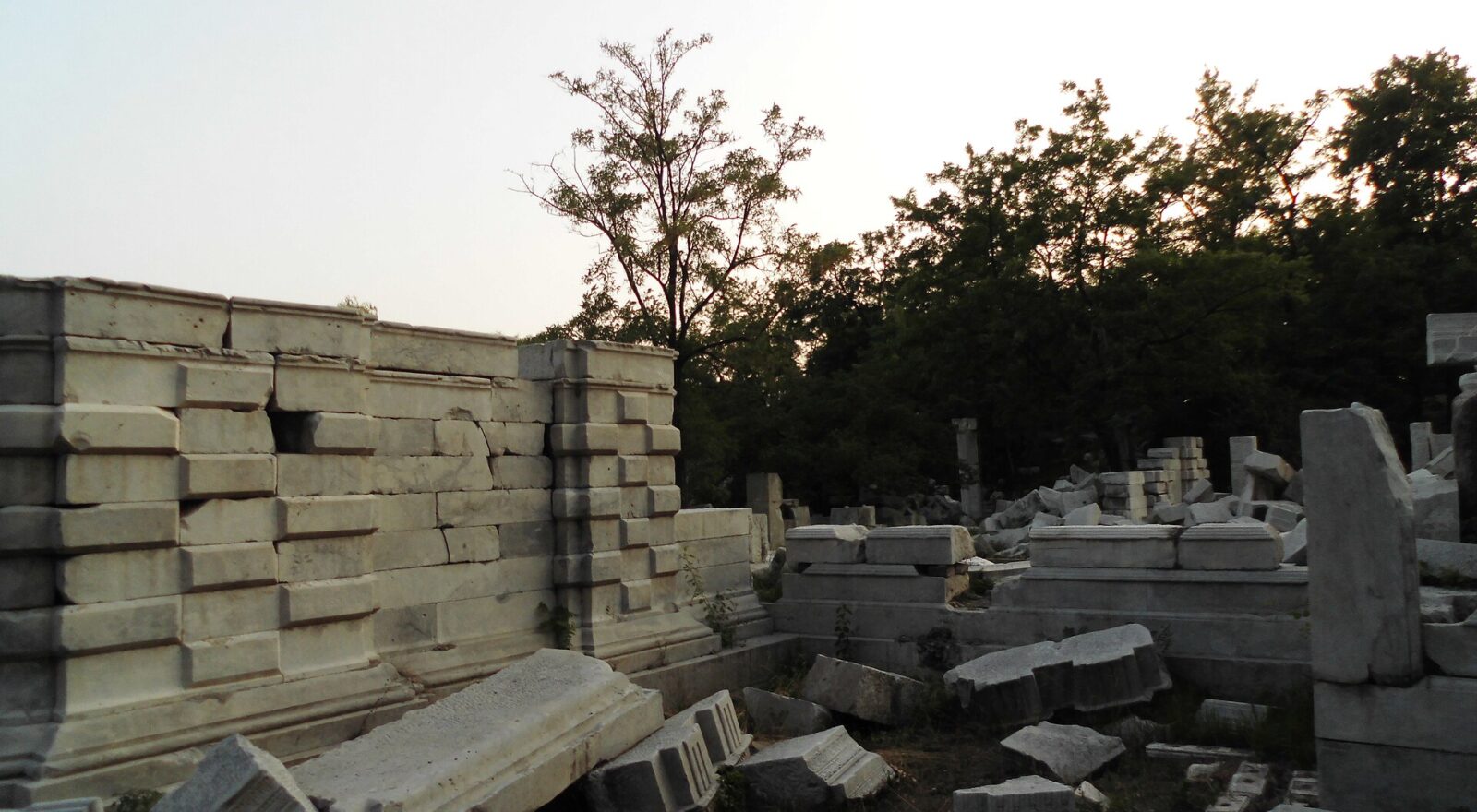 Carved white marble blocks and a broken wall among trees at the Western Mansions ruins in Yuanmingyuan