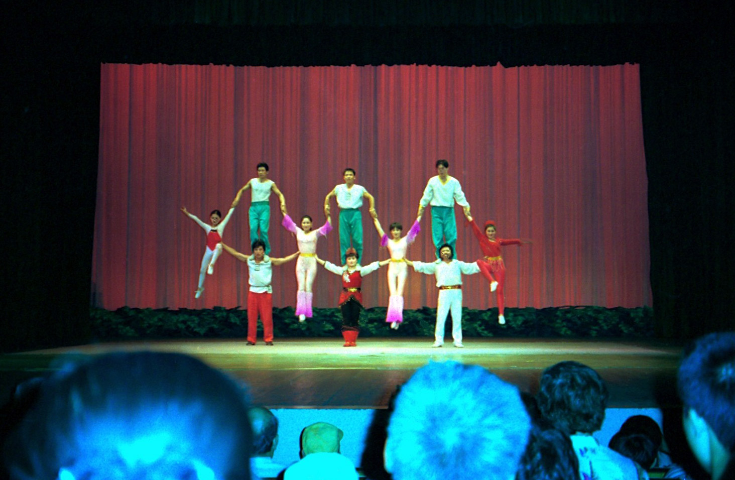 Acrobats in Beijing form a balanced group on stage before a pink curtain, with audience heads visible in the foreground.