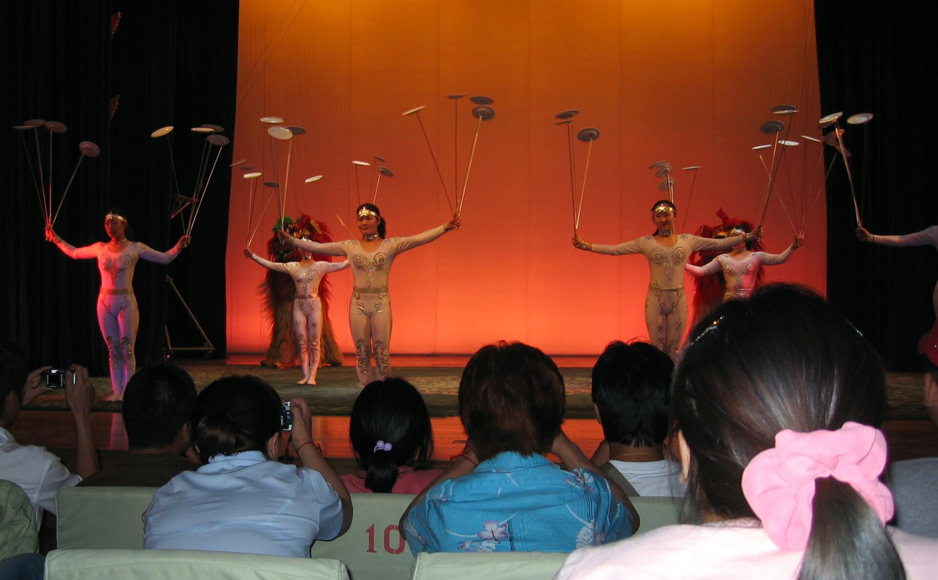 Acrobats on a stage spinning dozens of plates on tall poles in front of an orange curtain, watched by an audience in the foreground