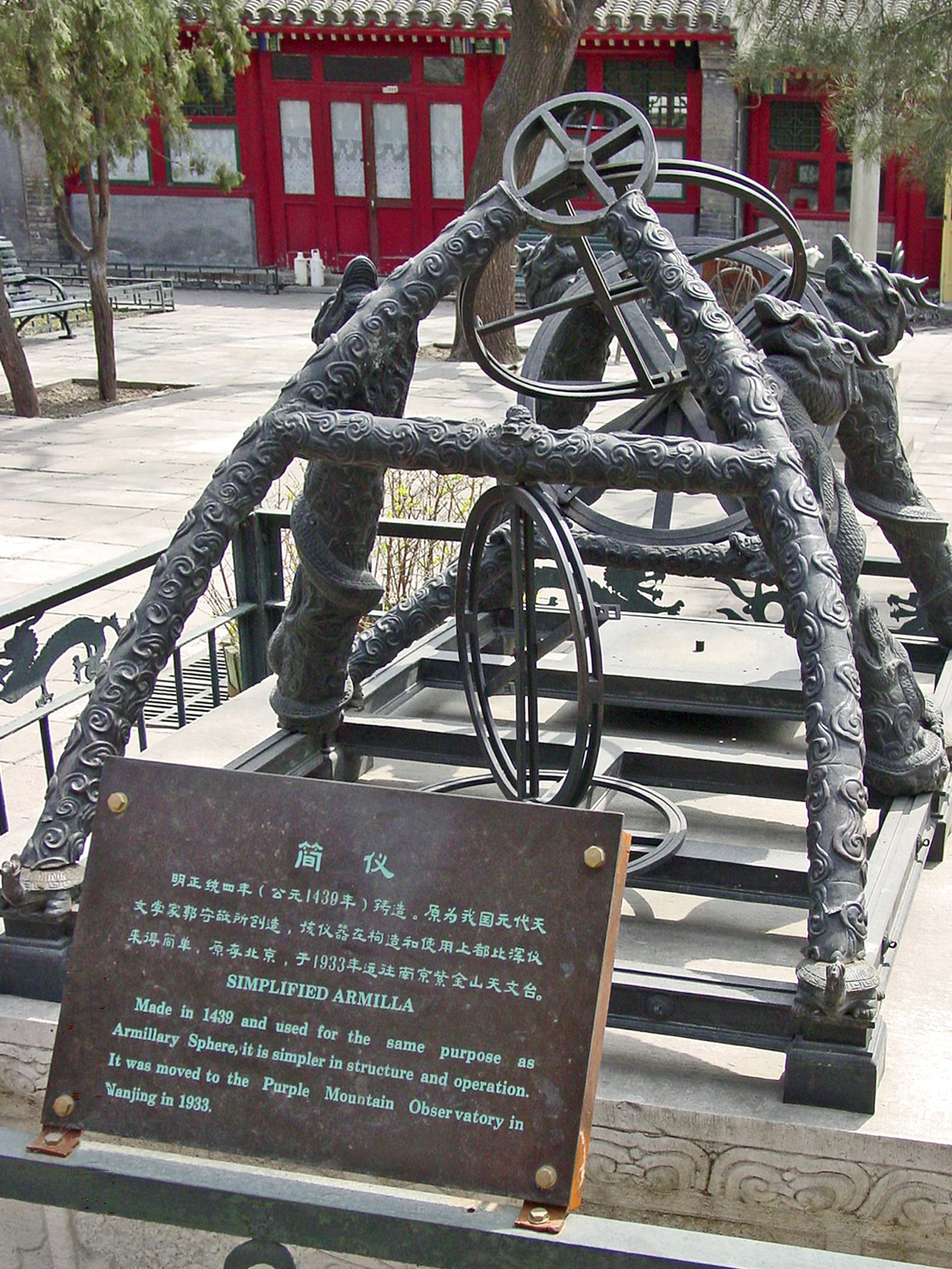 A bronze astronomical instrument on dragon-shaped supports in the courtyard of the Beijing Ancient Observatory, with a bilingual sign in front and a red-doored hall behind