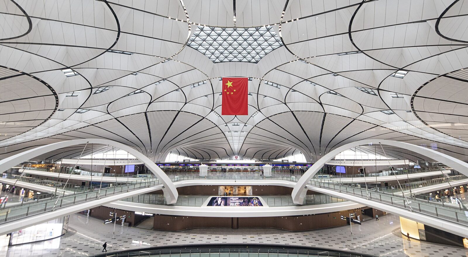 Beijing Daxing Airport has a sweeping white ceiling above several open terminal levels, with a Chinese flag hanging centrally.