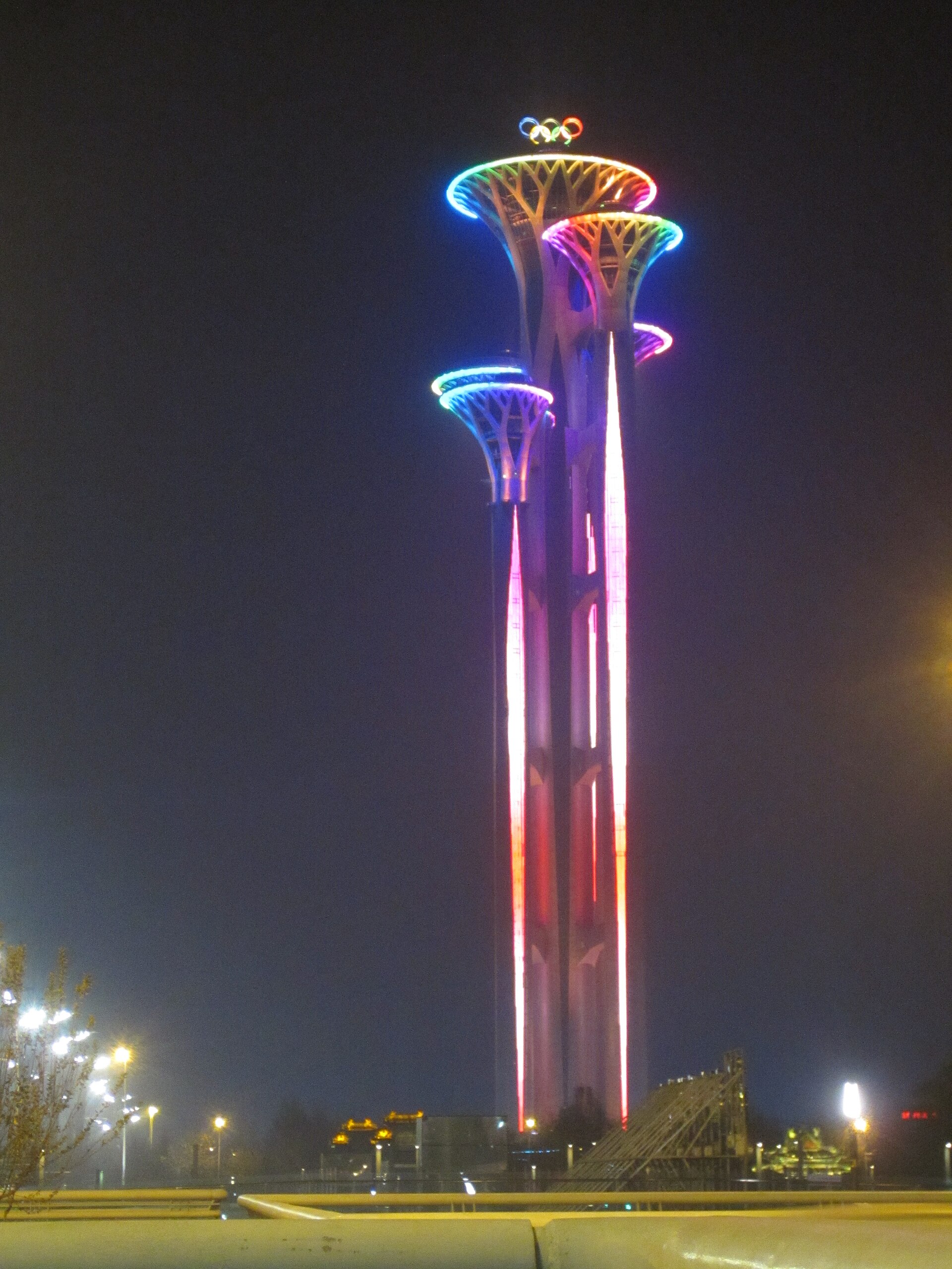 The Olympic Tower in Beijing at night, its five shafts lit in red and pink and its tree-like platforms ringed with coloured lights beneath the Olympic rings