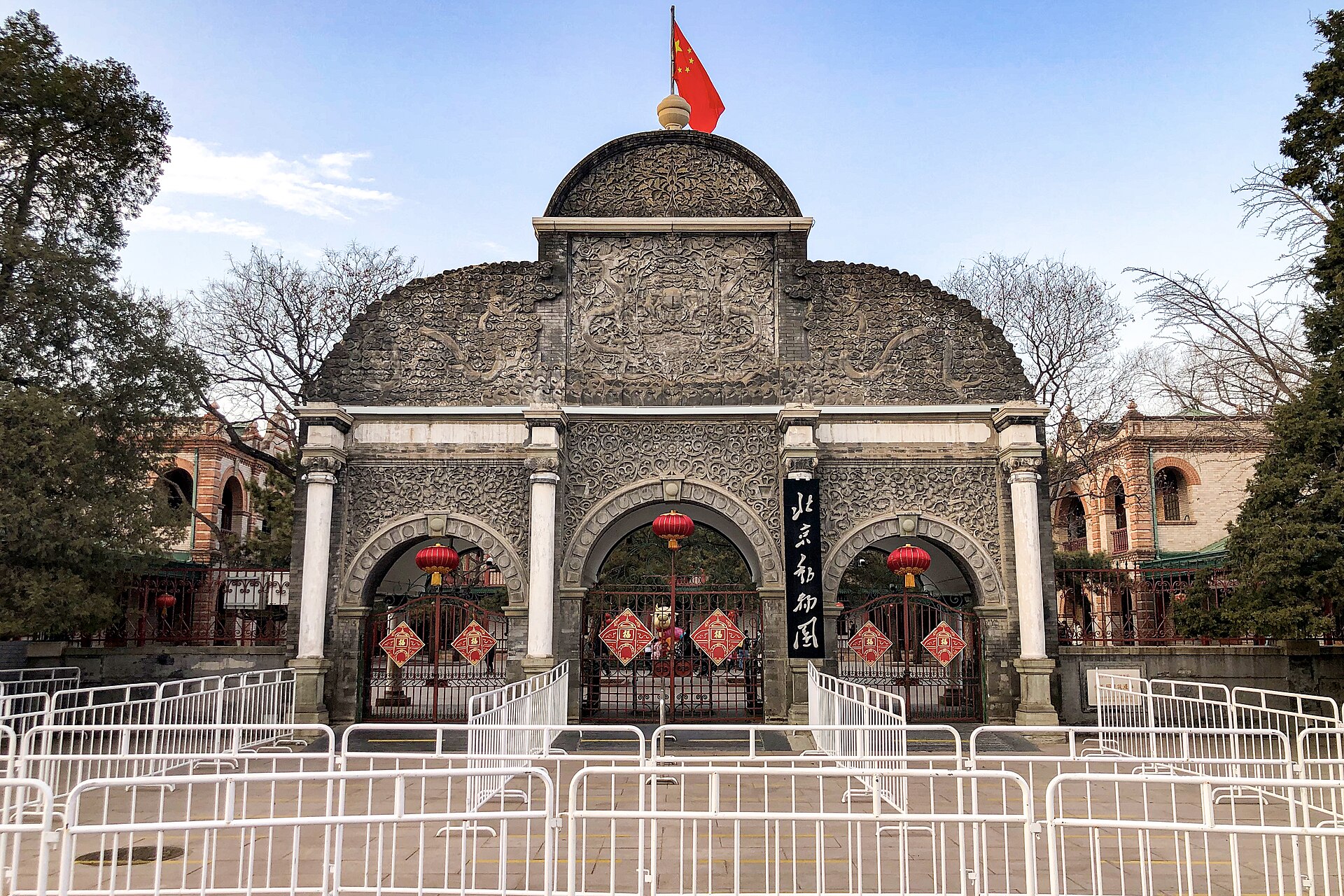 Beijing Zoo's carved stone front gate has three arches, red lanterns, a Chinese flag and white queue railings.