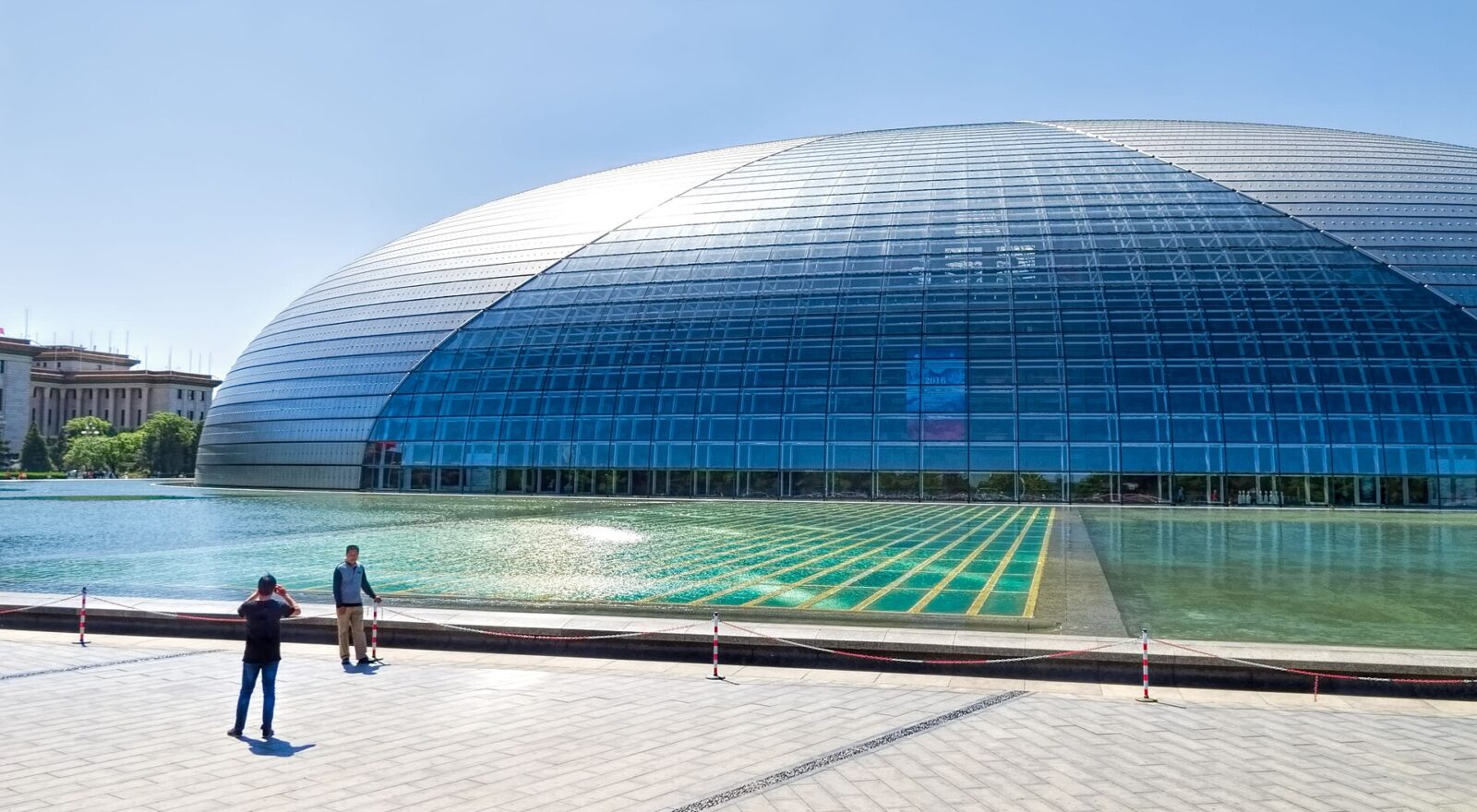 Beijing's National Centre for the Performing Arts curves above a reflecting pool as two visitors stand on the paved plaza.