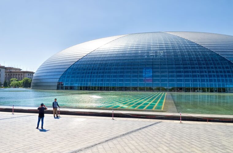 Beijing's National Centre for the Performing Arts curves above a reflecting pool as two visitors stand on the paved plaza.