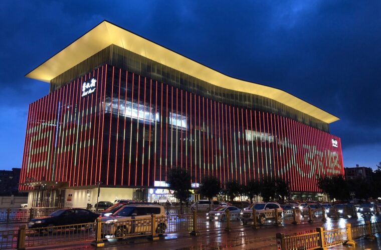 Beijing's Silk Market glows with red vertical lights and a golden roofline above traffic on a wet street at dusk.