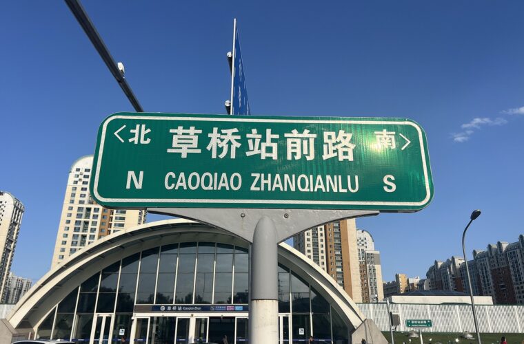 A bilingual Caoqiao Zhanqianlu street sign stands in front of Caoqiao subway station in Beijing under a blue sky.