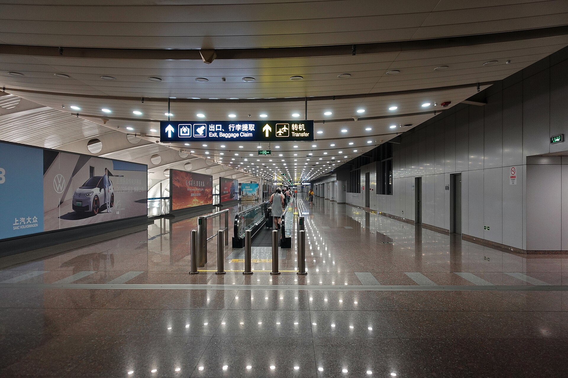 Bilingual exit, baggage claim and transfer signs hang above a moving walkway in Beijing Capital Airport Terminal 2 arrivals.
