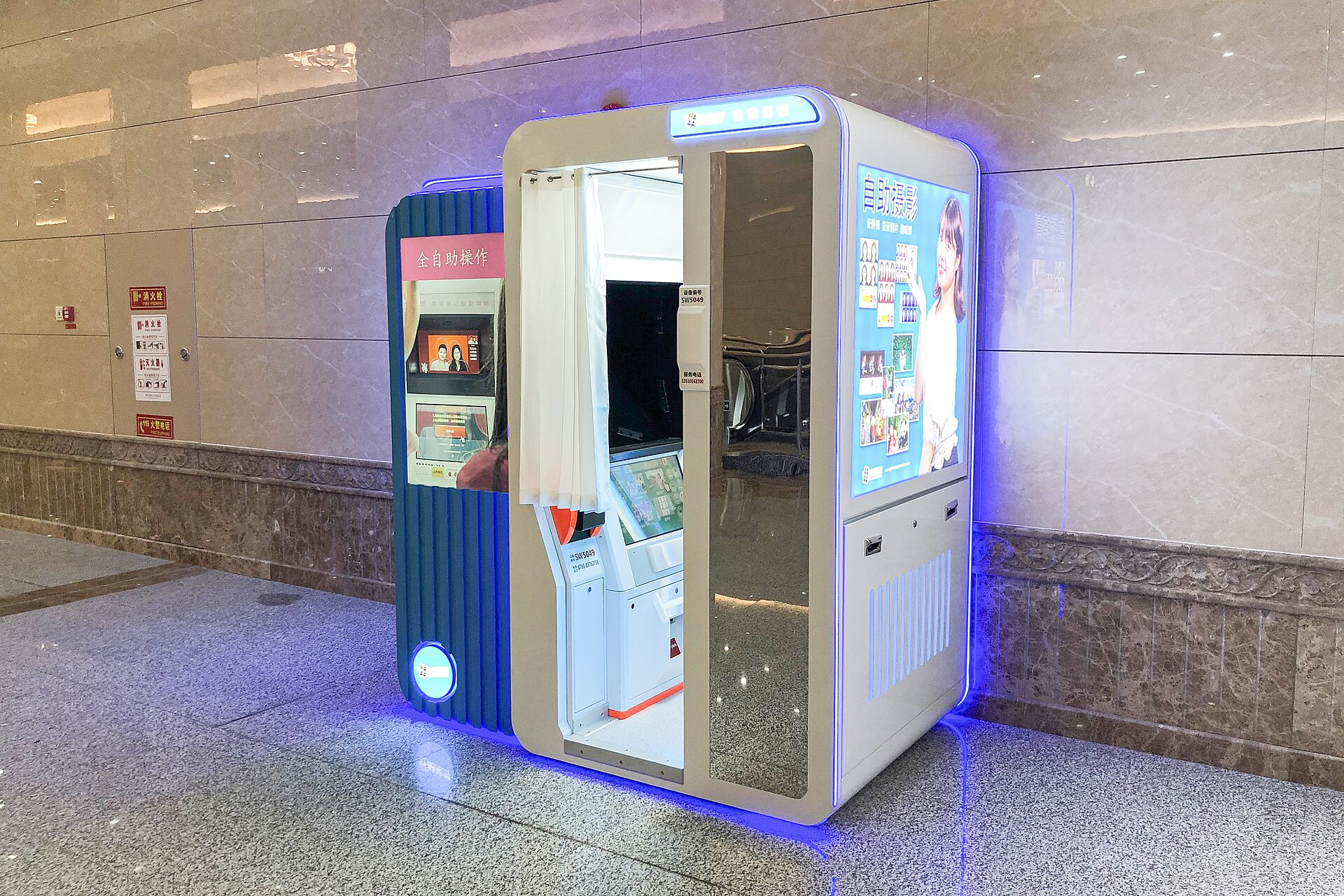 A blue-and-white self-service photo booth with a white curtain and touchscreen stands inside Beijing's Ganjiakou subway station.