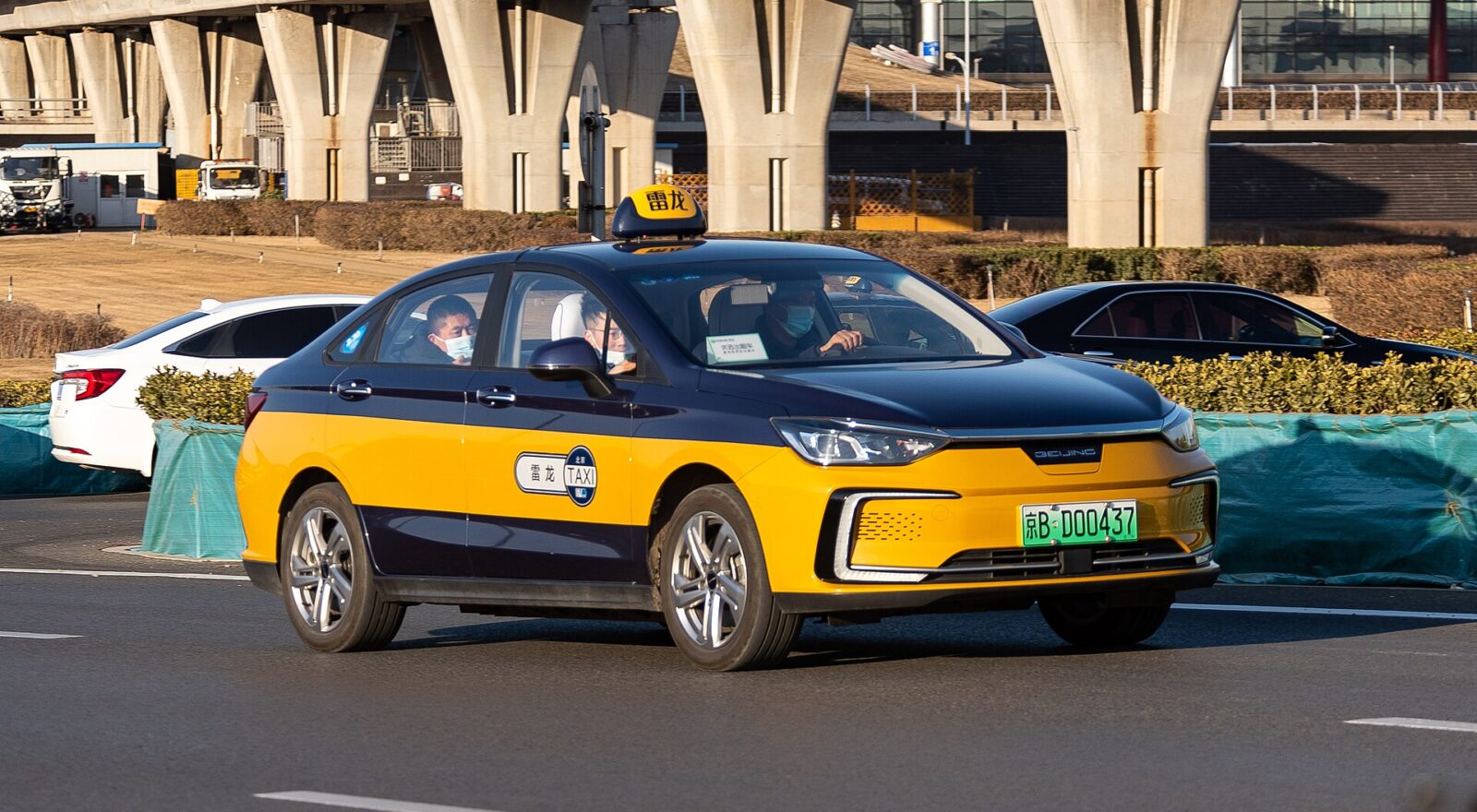 Blue and yellow Beijing taxi passes elevated airport tracks, with a roof sign and green Beijing registration plate.