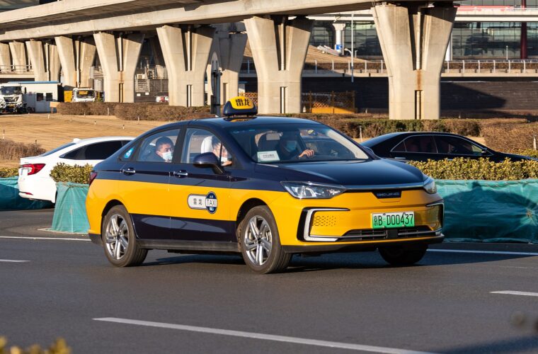 Blue and yellow Beijing taxi passes elevated airport tracks, with a roof sign and green Beijing registration plate.