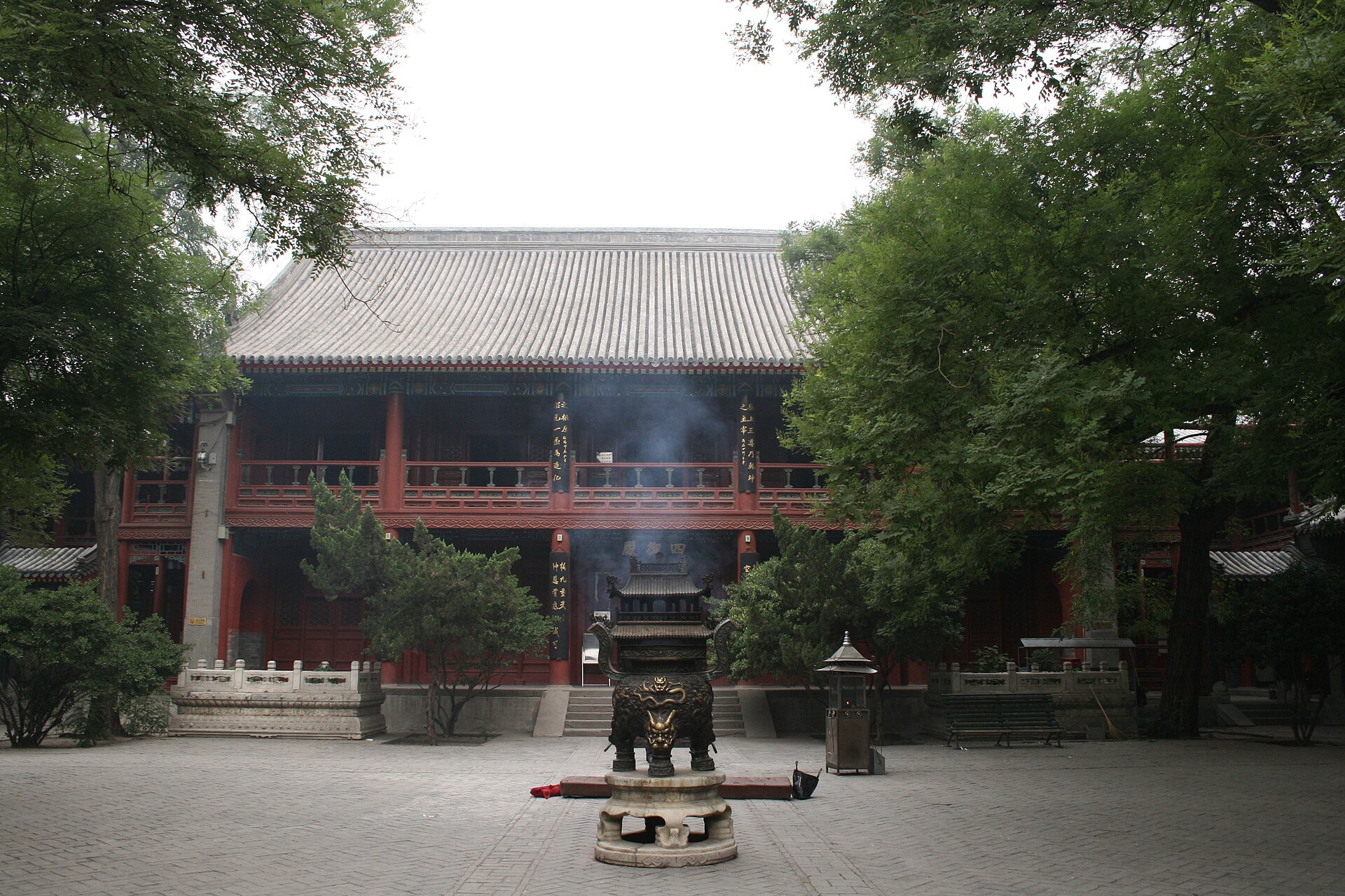 Bronze incense burner with rising smoke in a paved courtyard facing a red-pillared hall with a grey tiled roof at the White Cloud Temple, Beijing