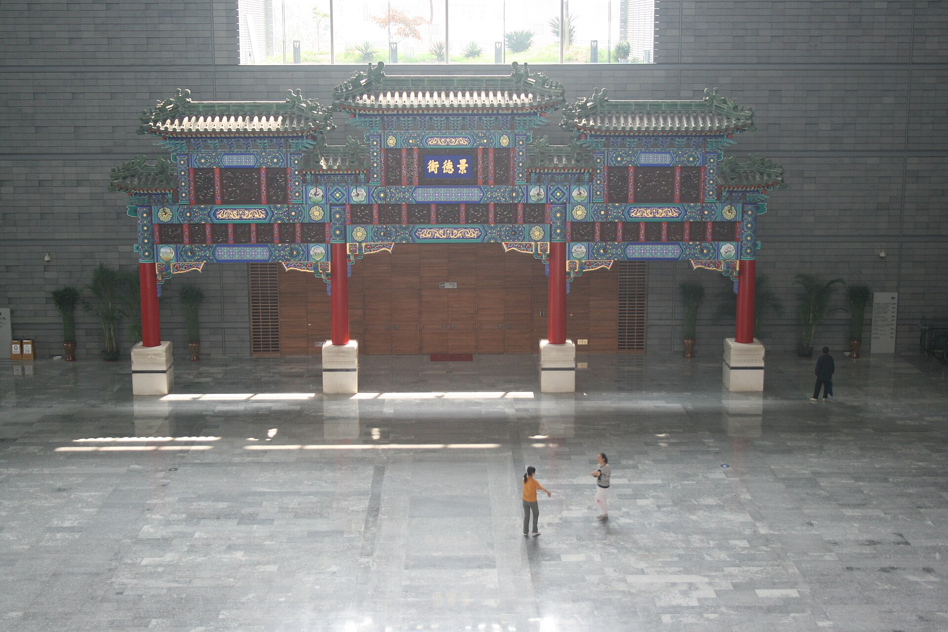 A painted memorial archway with green-tiled roofs, red columns and a blue panel reading Jingde Street, standing in the stone-floored atrium of the Capital Museum