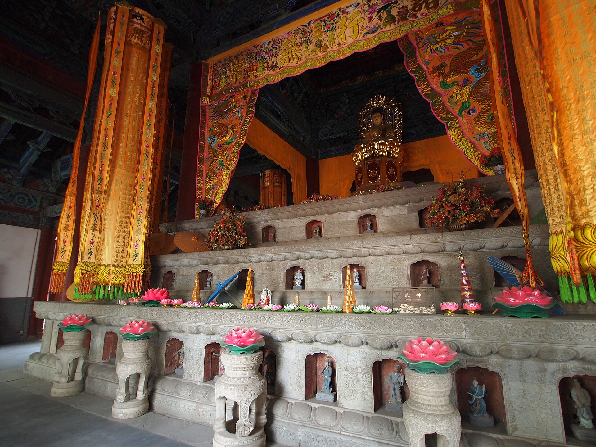 Carved figures line the stone tiers of Jietai Temple's Ordination Terrace beneath colorful embroidered hangings and a gilded figure.
