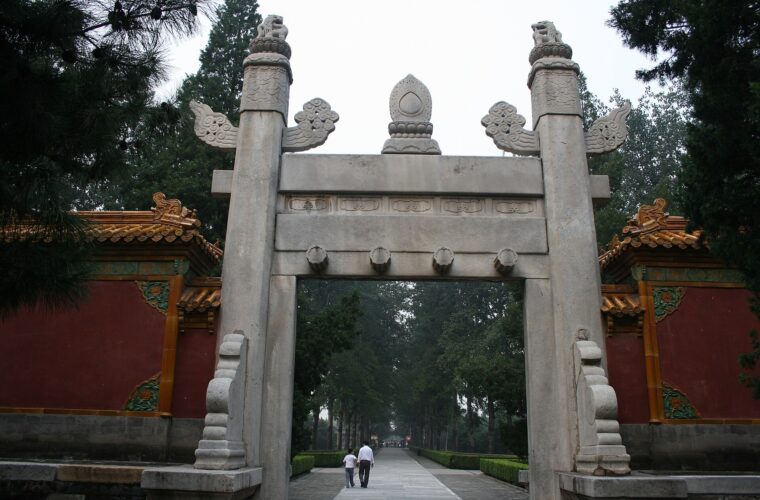 A carved stone archway between red walls frames visitors and the tree-lined Sacred Way at Beijing's Ming Tombs.