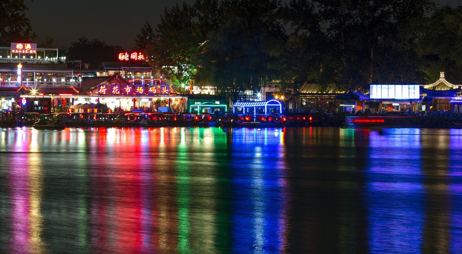 Colorful lights from lakeside buildings and boats reflect across dark Houhai water in Beijing beneath silhouetted trees at night.