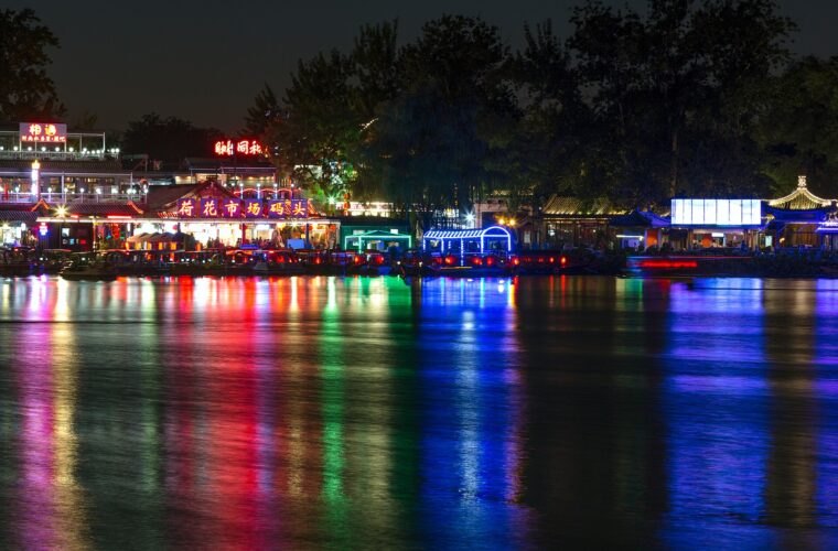 Colorful lights from lakeside buildings and boats reflect across dark Houhai water in Beijing beneath silhouetted trees at night.
