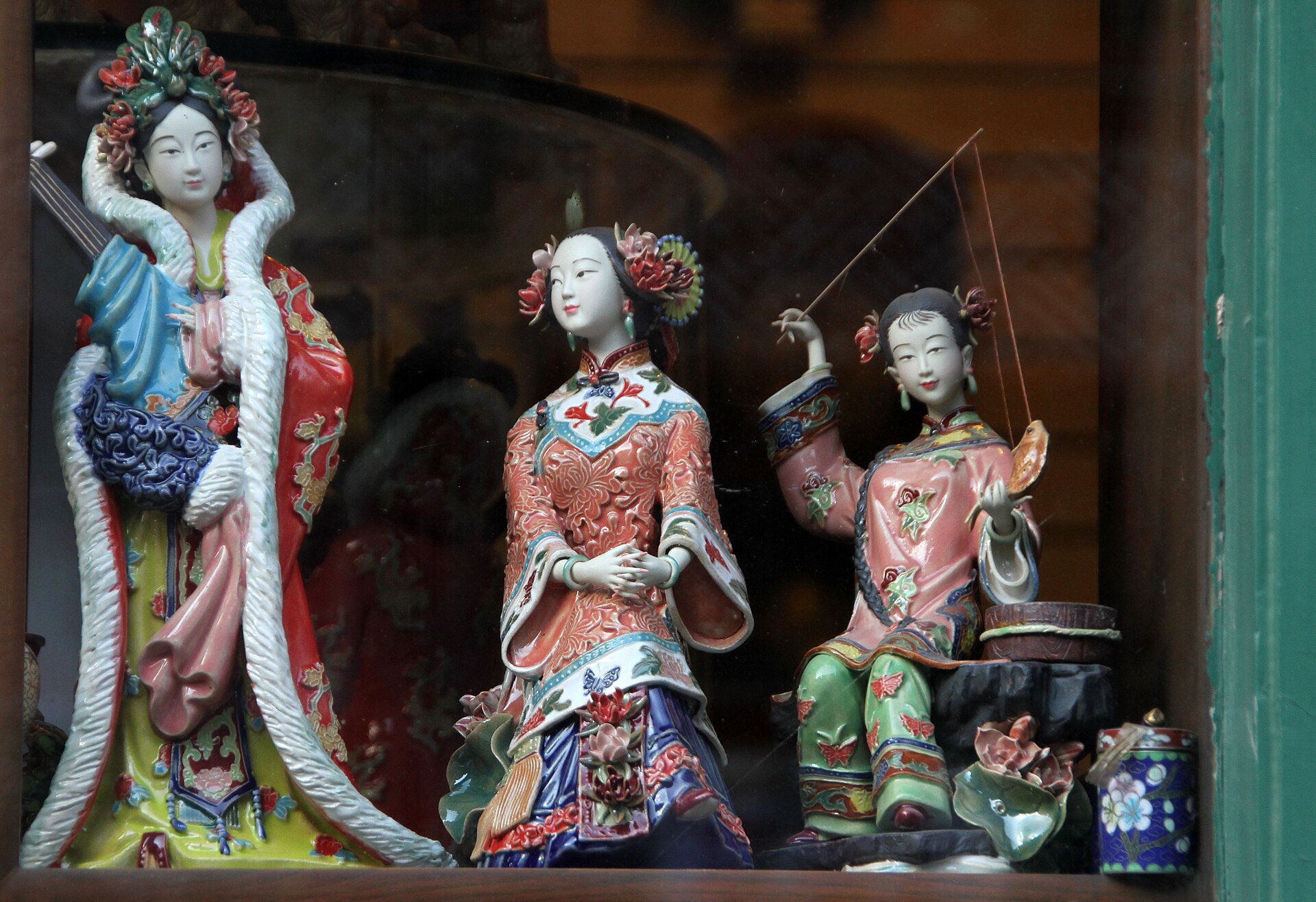 Colorful porcelain figurines wearing ornate robes stand in a shop window on Beijing Liulichang, including one holding a fan.