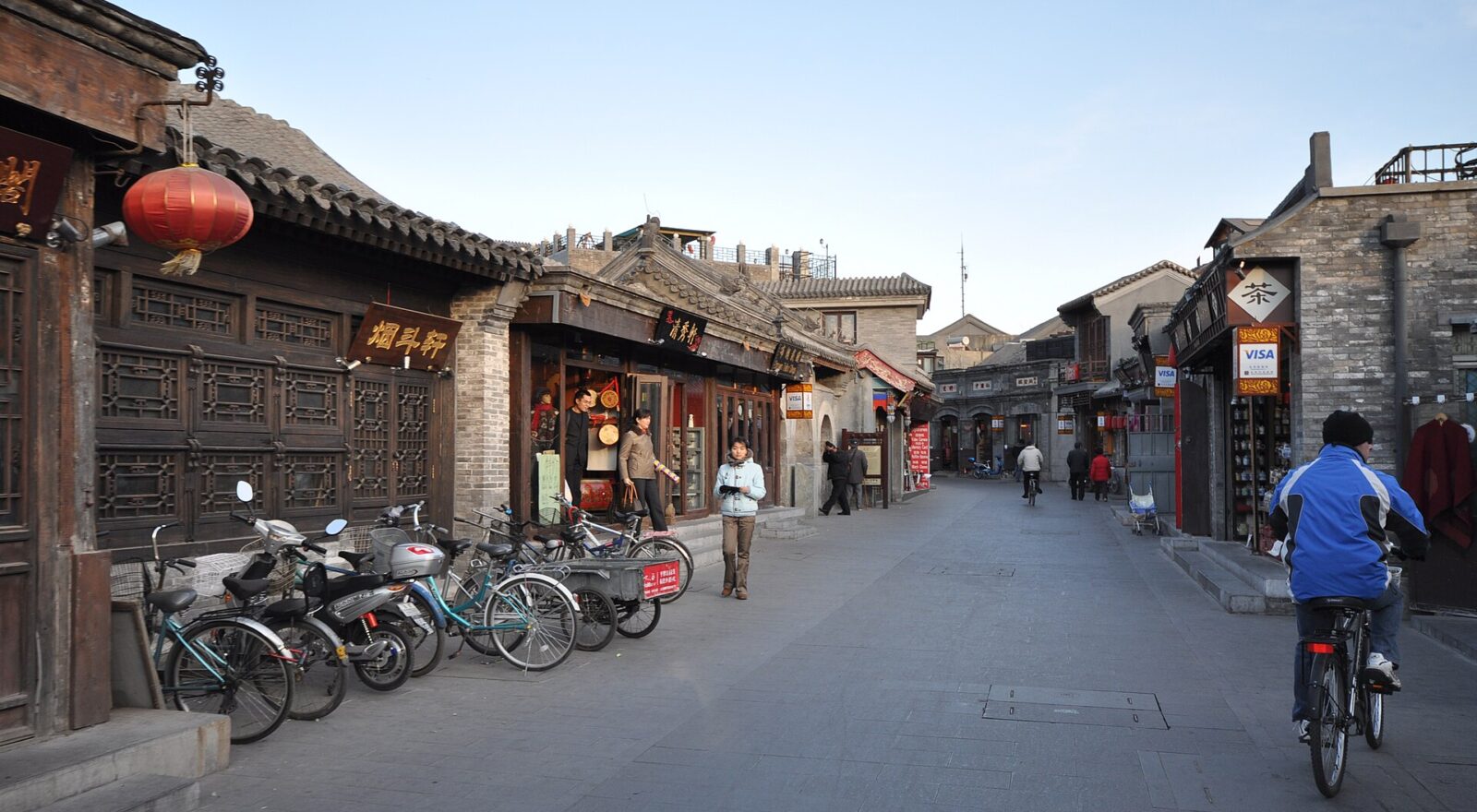 A cyclist in a blue jacket rides along Beijing's Yandai Xiejie past traditional shopfronts, parked bicycles and pedestrians.