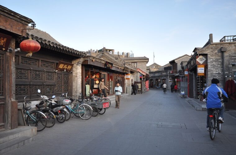 A cyclist in a blue jacket rides along Beijing's Yandai Xiejie past traditional shopfronts, parked bicycles and pedestrians.