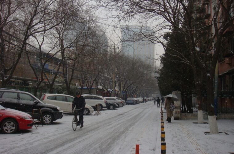A cyclist and umbrella-carrying pedestrian travel along a snowy Beijing street lined with parked cars and bare winter trees.