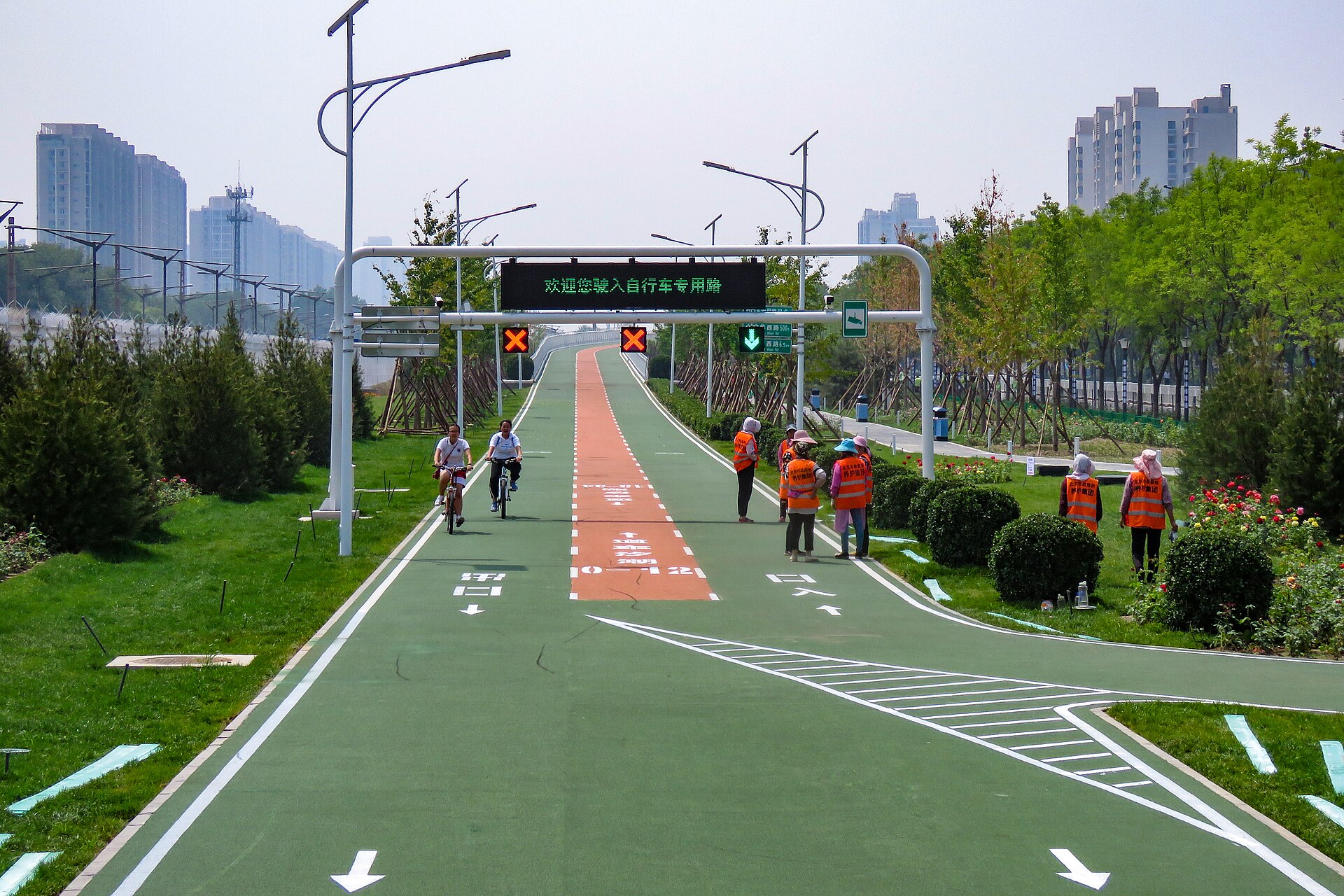 Cyclists ride along Beijing's Huilongguan-Shangdi Bicycle Lane beneath overhead signals, with marked lanes and landscaped verges.