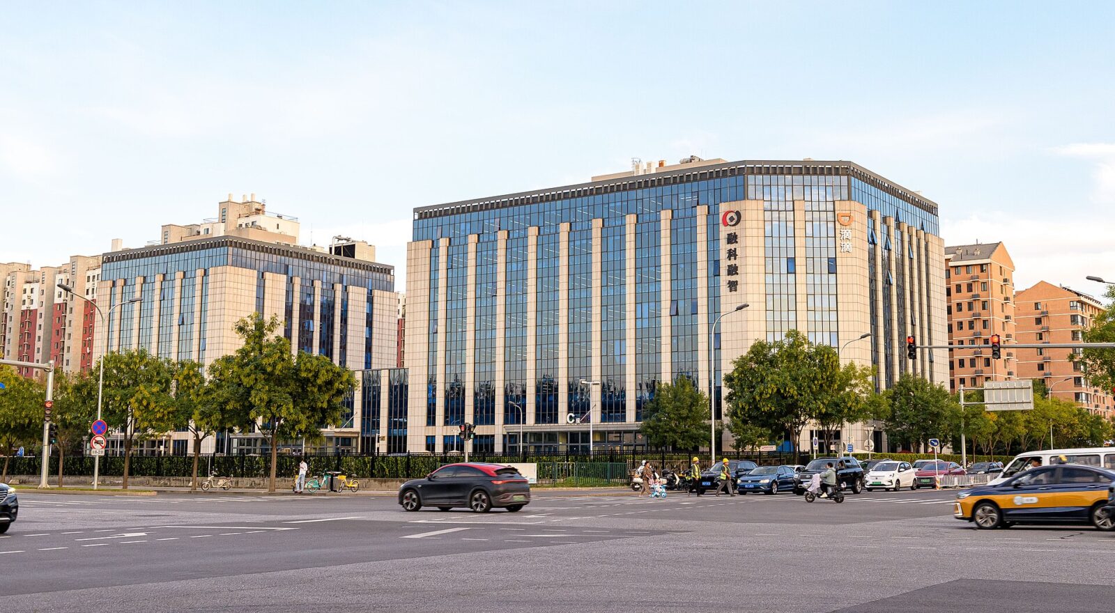 DiDi's headquarters, a large glass office building with the DiDi logo on its corner, seen across a Beijing street where a yellow taxi and other cars are passing