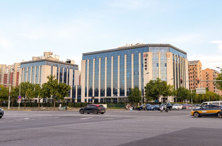 DiDi's headquarters, a large glass office building with the DiDi logo on its corner, seen across a Beijing street where a yellow taxi and other cars are passing