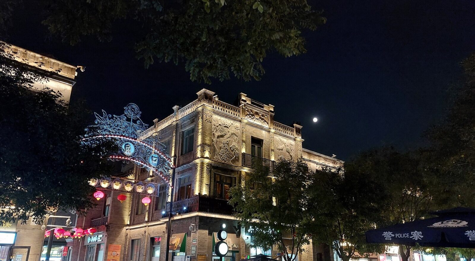 An evening crowd passes illuminated shopfronts and red lanterns on Qianmen Dajie in Beijing beneath leafy trees and the moon.
