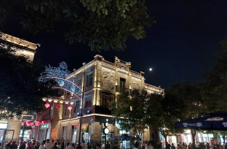 An evening crowd passes illuminated shopfronts and red lanterns on Qianmen Dajie in Beijing beneath leafy trees and the moon.