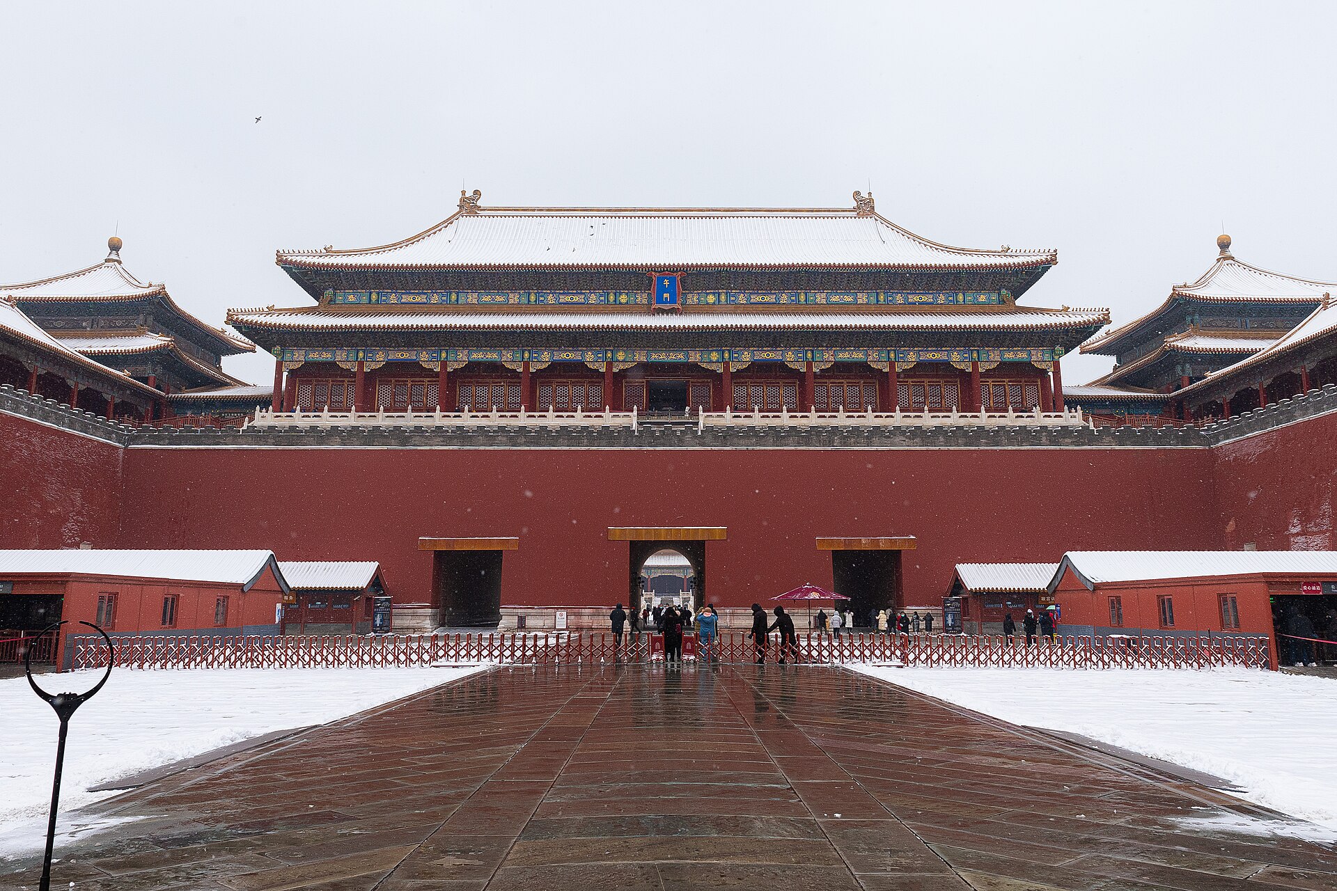 The Meridian Gate of the Forbidden City in falling snow, with red walls, snow-covered roofs and visitors passing through the ticket check barriers in front