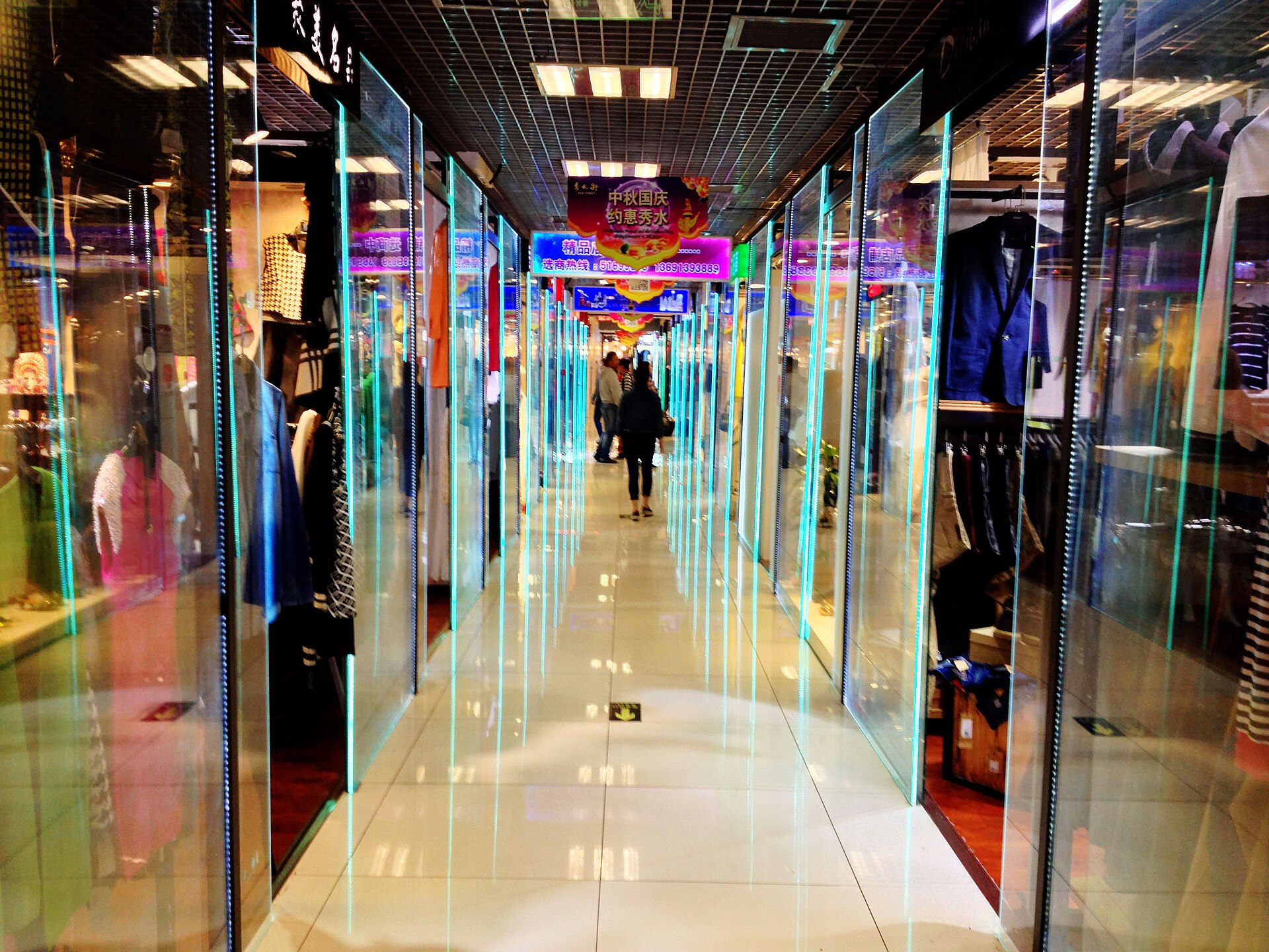 Glass-fronted clothing shops and mannequins line a bright tiled aisle inside Beijing's Silk Market, with shoppers farther along.