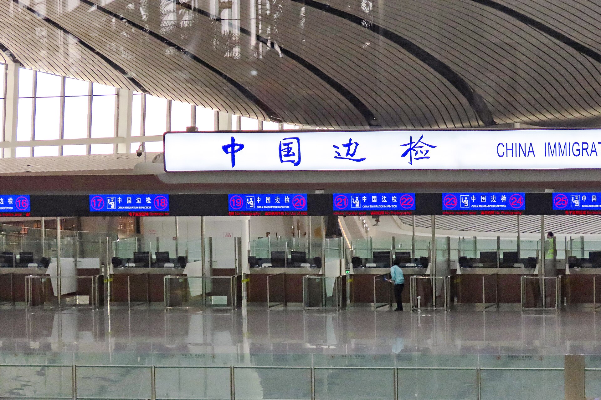 Glass-fronted immigration counters stand beneath illuminated Chinese and English border-inspection signs inside Beijing Daxing International Airport.