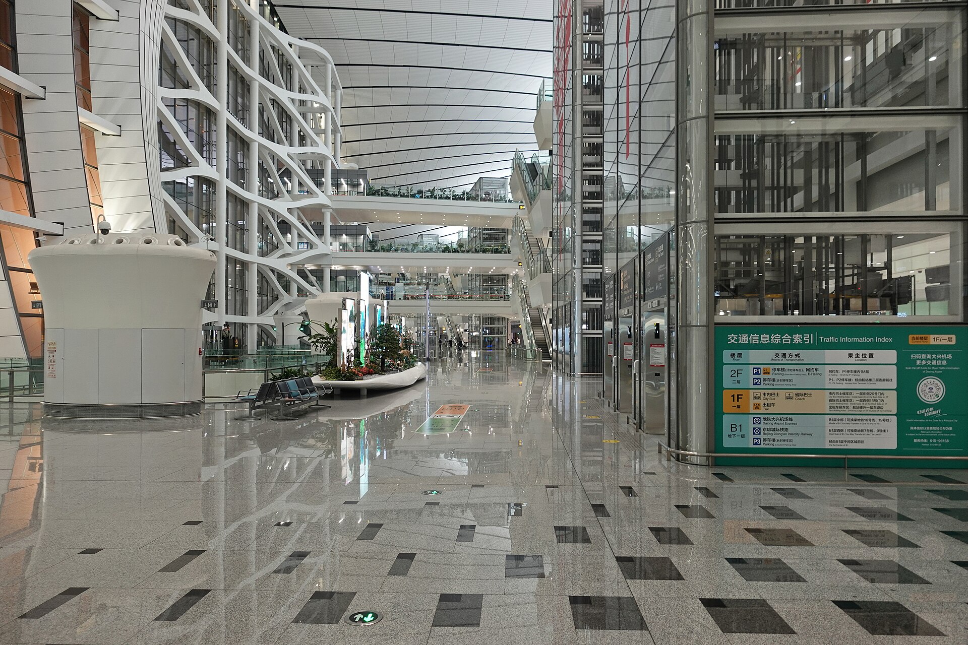 Glass lifts and elevated walkways rise above Beijing Daxing Airport B1 hall, beside a green bilingual transport information directory.