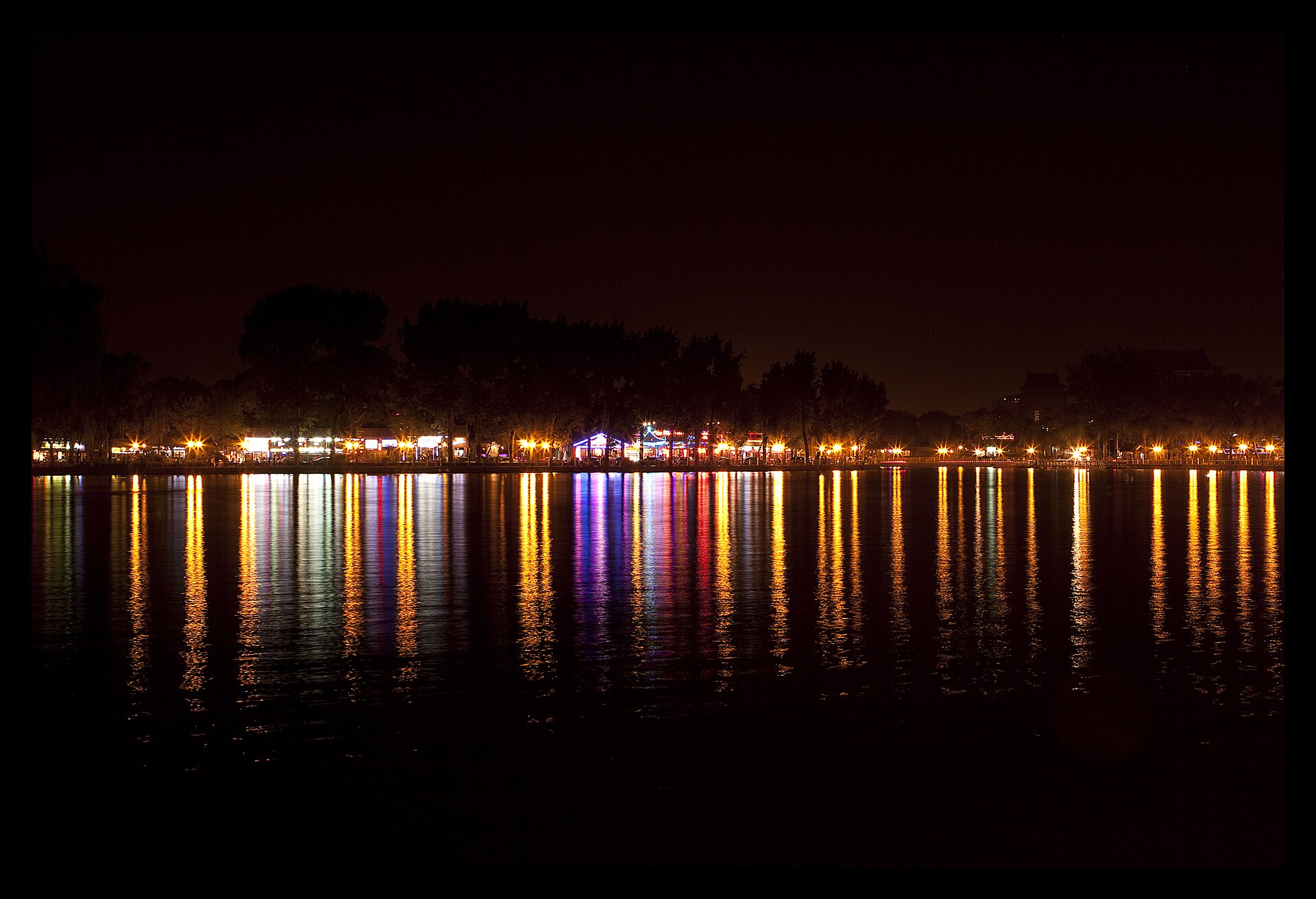 Gold and purple bar lights reflect across Beijing Houhai Lake beneath a dark shoreline of trees at night.