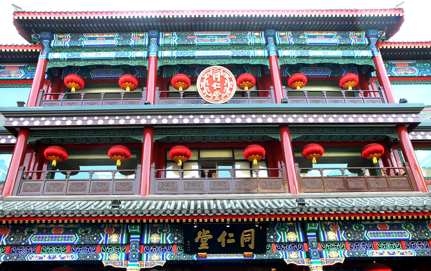 Gold Tongrentang lettering stands above the entrance beneath red columns, lanterns and richly painted eaves in Beijing's Dashilar area.