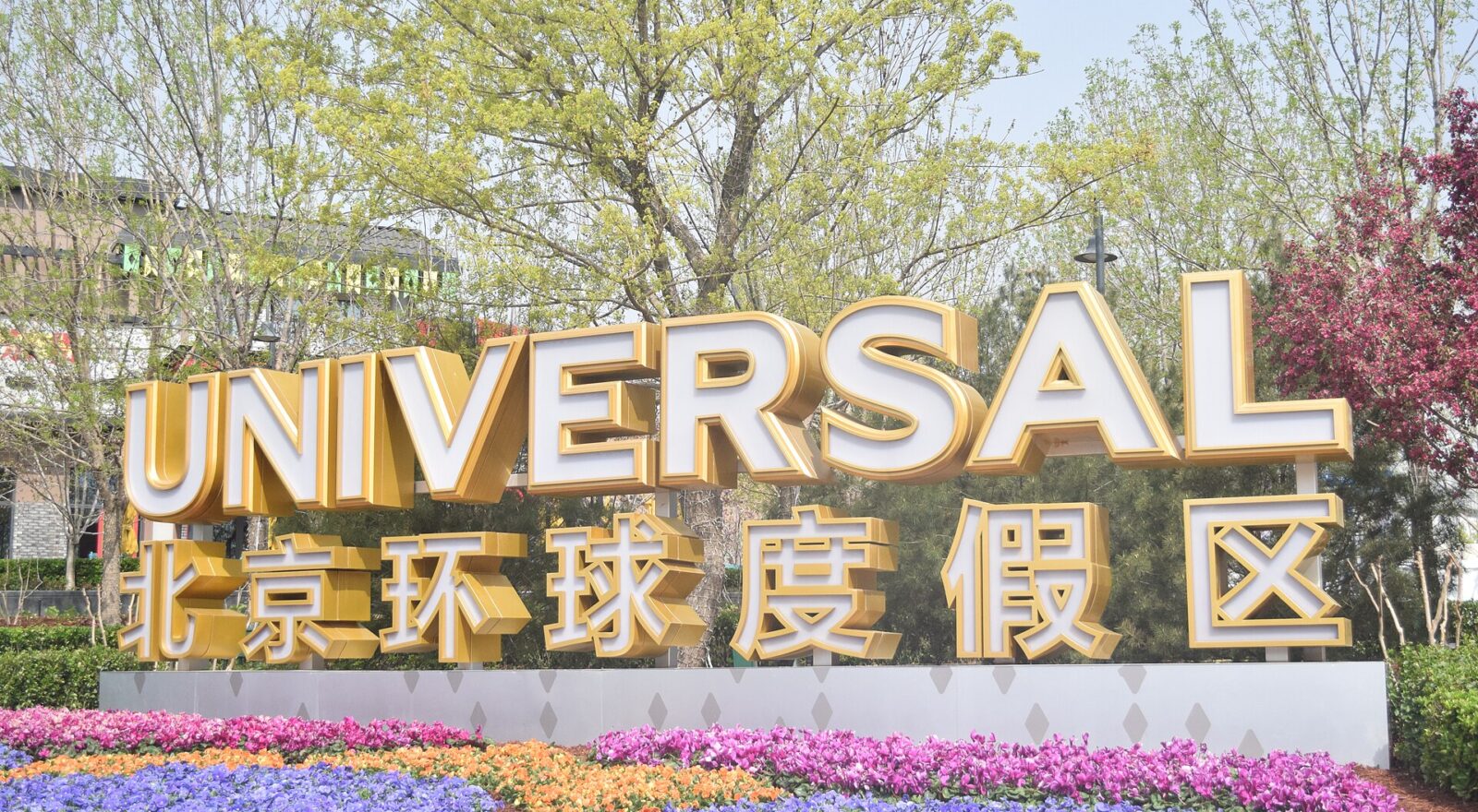 Gold Universal Beijing Resort entrance lettering rises above colourful flower beds, with leafy trees behind the bilingual sign.