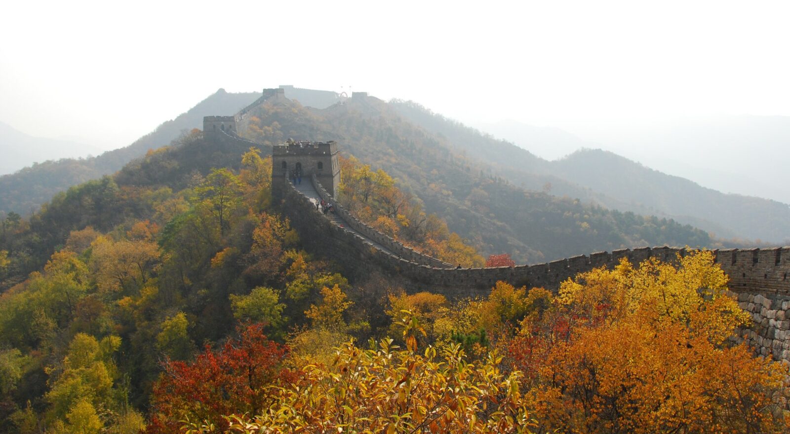 Great Wall of China winding along a ridge past a watchtower, with yellow, orange and red autumn trees in the foreground and hazy mountains behind