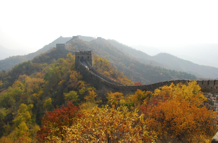 Great Wall of China winding along a ridge past a watchtower, with yellow, orange and red autumn trees in the foreground and hazy mountains behind