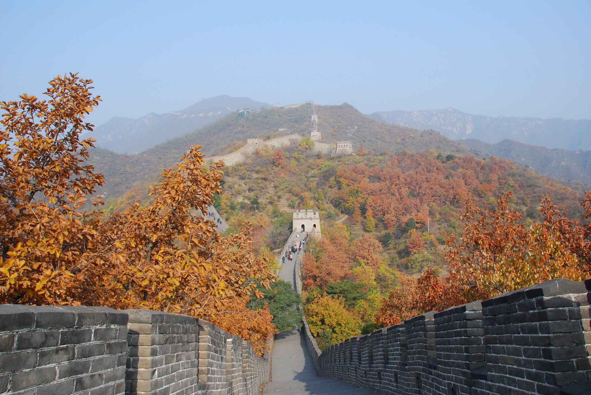 Great Wall path between brick parapets with watchtowers on the ridge ahead, orange autumn leaves in the foreground and red-orange hillsides beyond