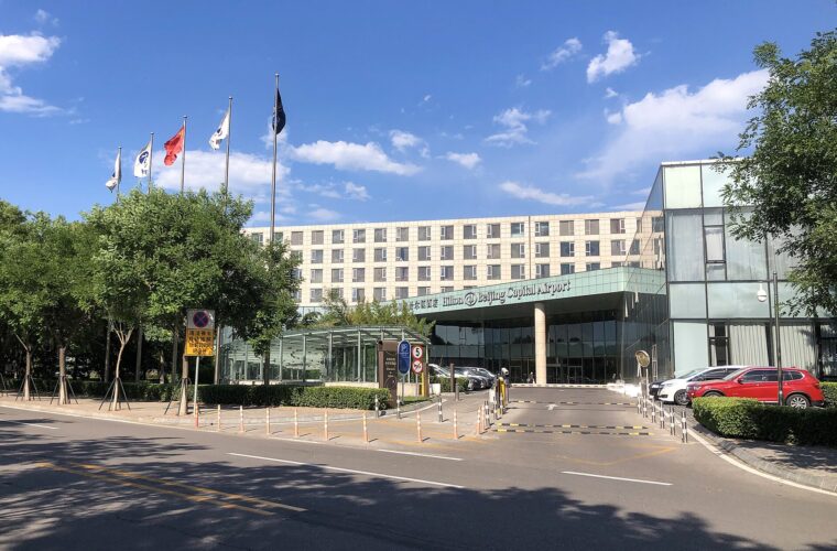 Hilton Beijing Capital Airport's entrance canopy and hotel wing face a driveway bordered by trees and flagpoles under blue skies.