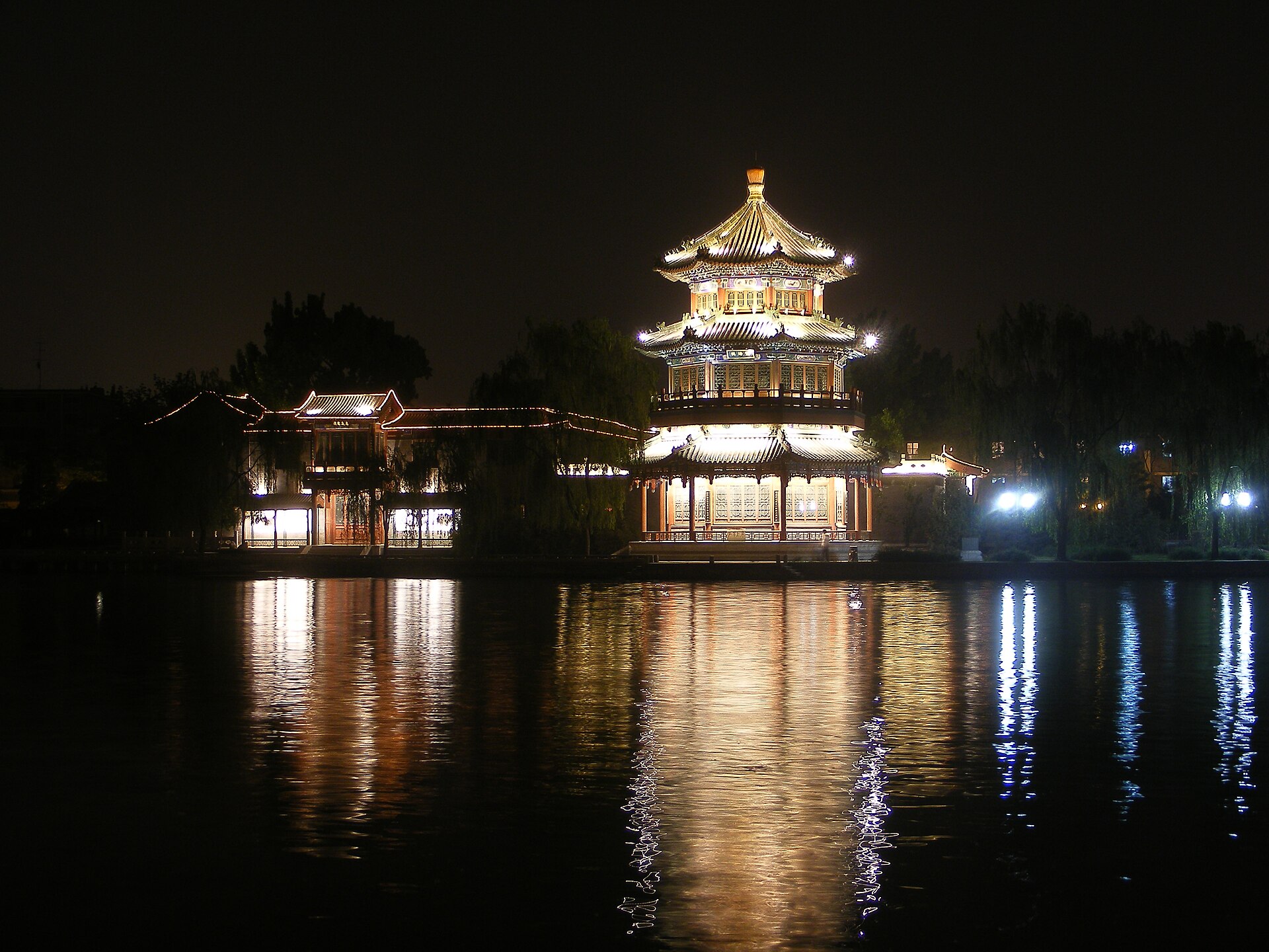 An illuminated tiered pavilion and nearby buildings cast gold reflections across the dark Shichahai water in Beijing at night.