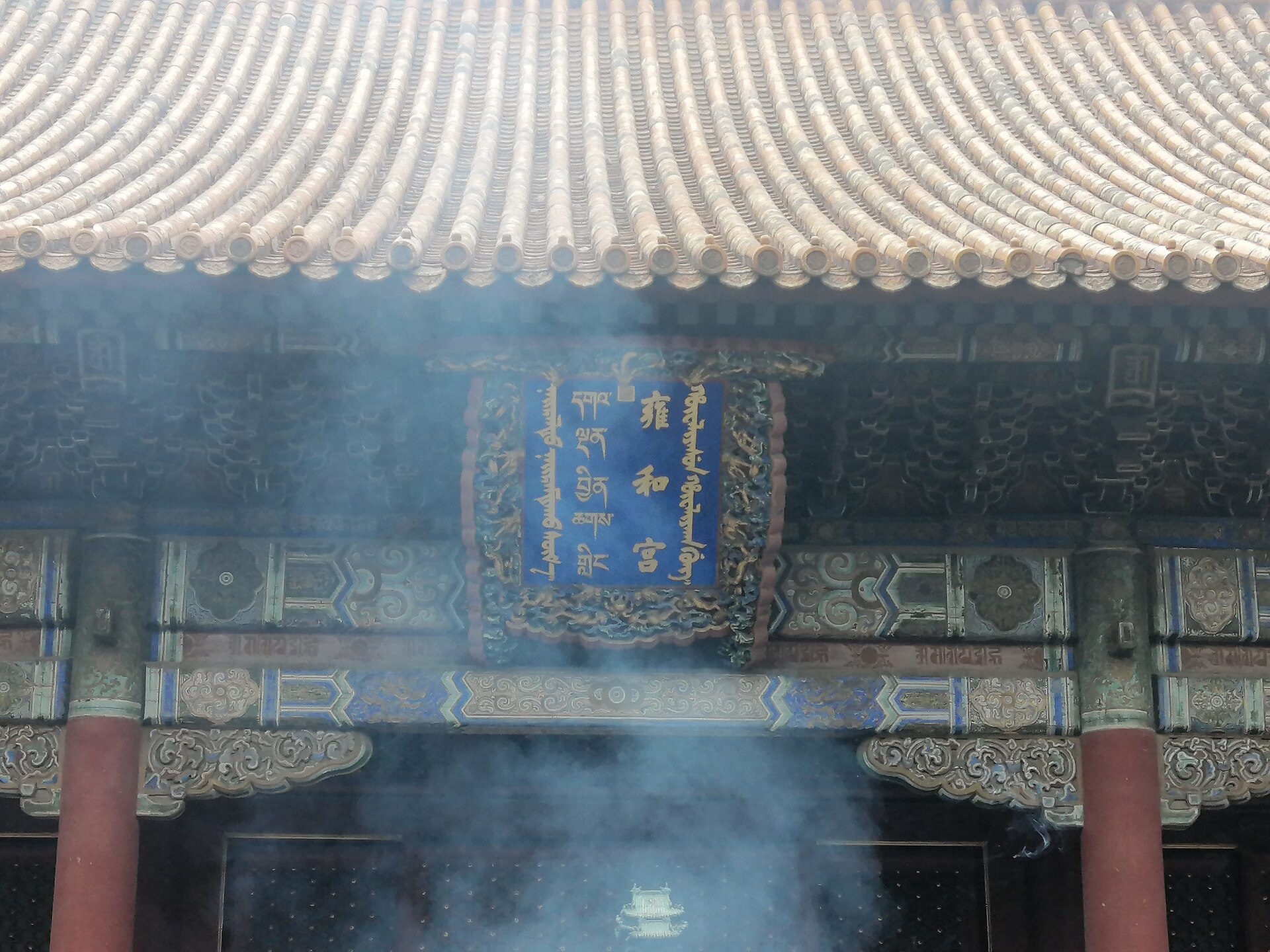 Incense smoke drifts across red columns and the blue quadrilingual name plaque beneath yellow roof tiles at Beijing's Lama Temple.