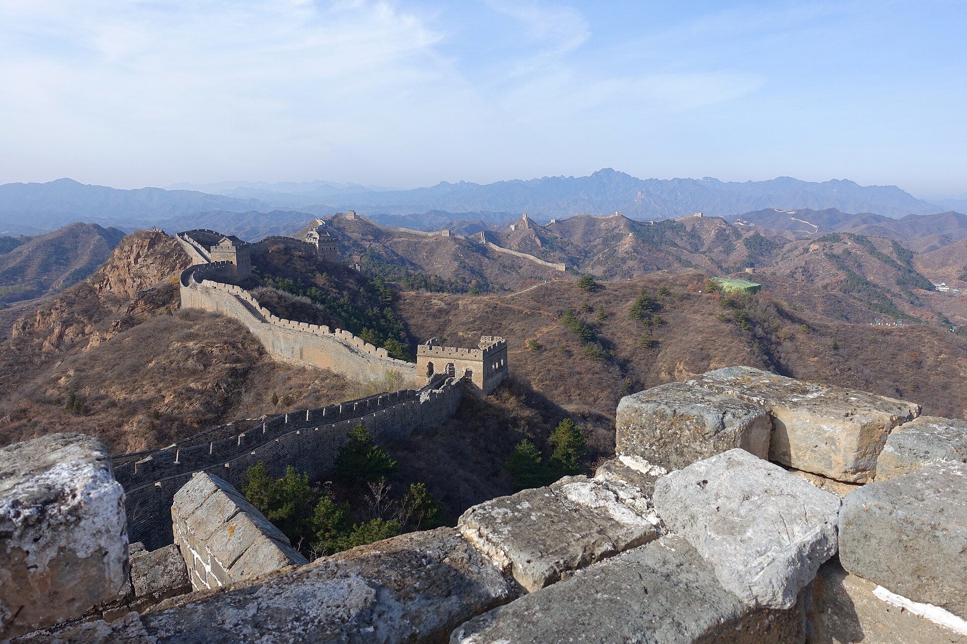 The Great Wall at Jinshanling running along a ridge of brown autumn hills with watchtowers, seen from a crumbling stone parapet in the foreground