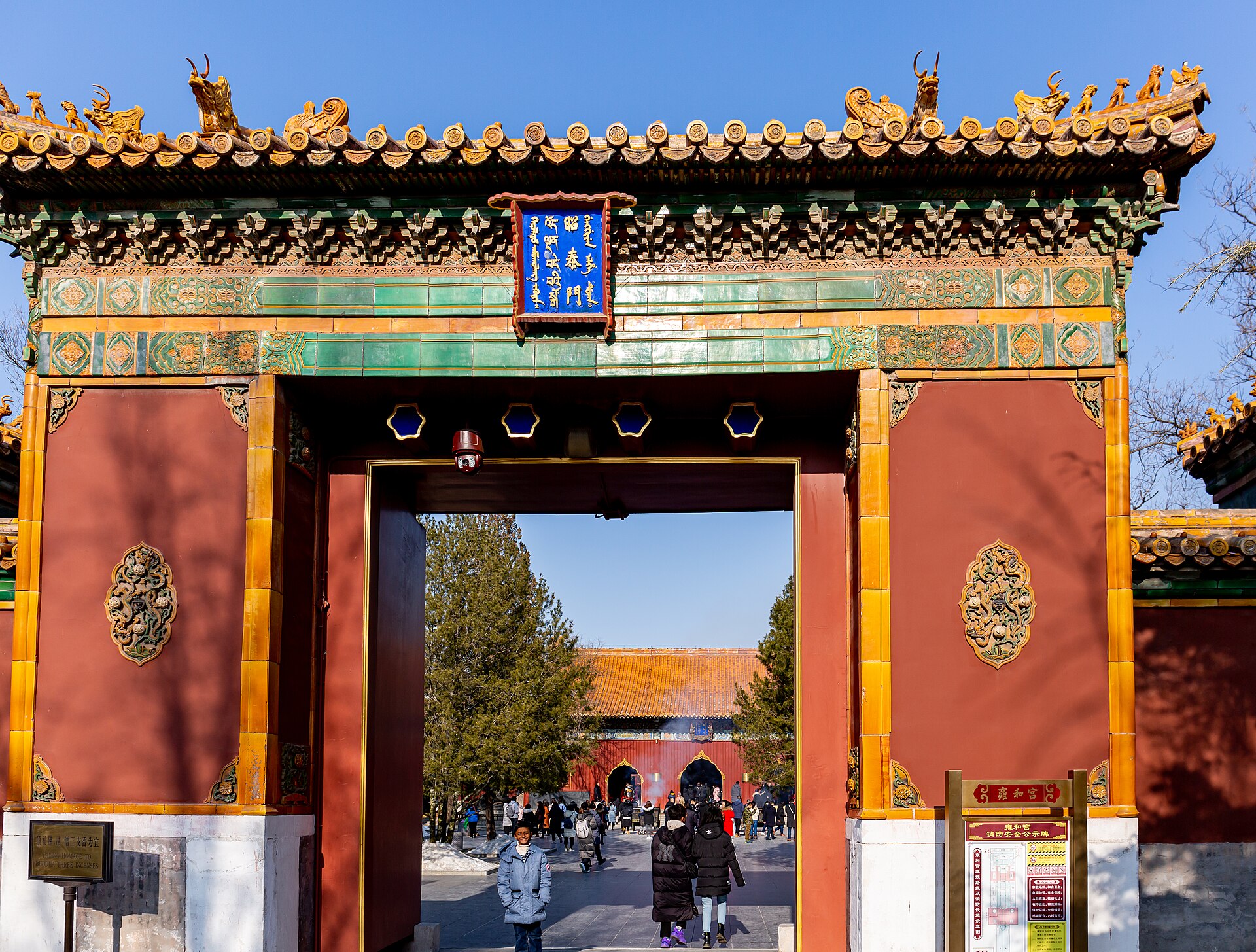 Visitors walking through a red gate with yellow glazed tiles and a blue plaque into the courtyards of the Lama Temple in Beijing on a clear winter day